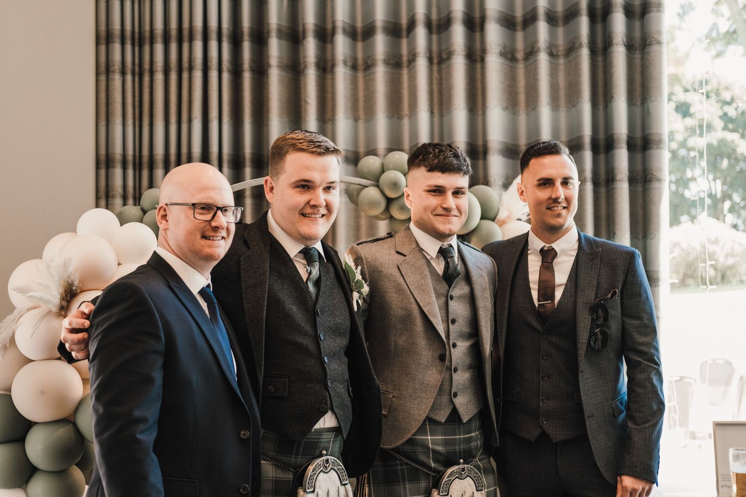 Four groomsmen in formal attire stand together smiling at a wedding with balloon decor in the background.