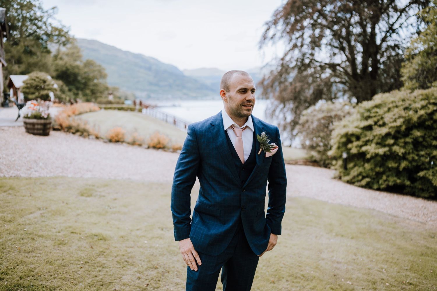 Person in navy suit standing by lake with mountains in background.