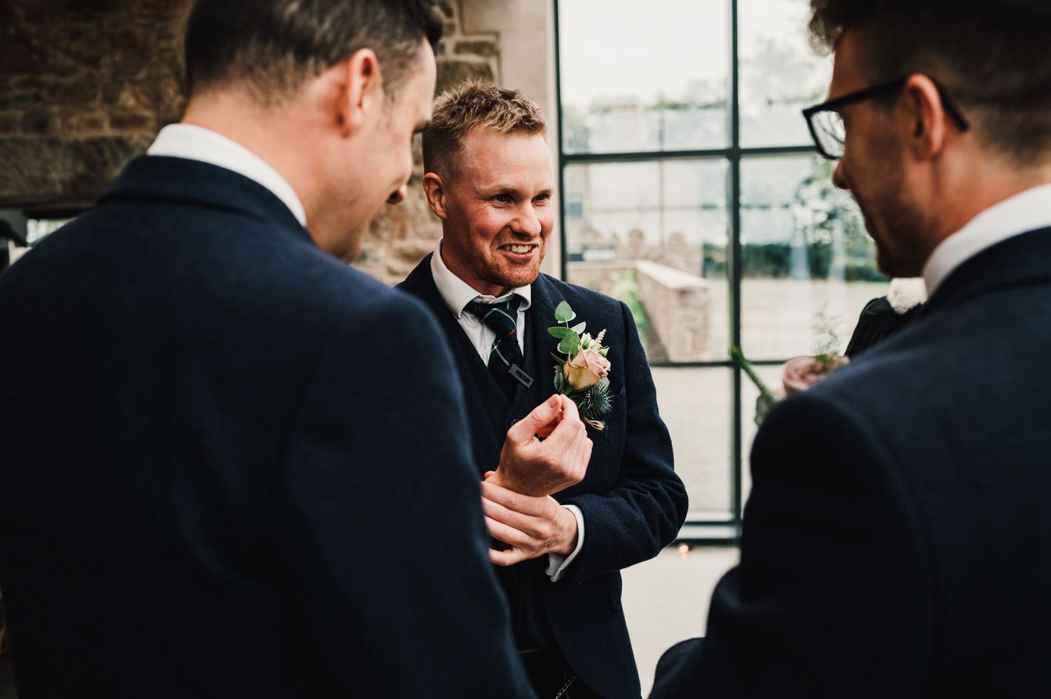Candid moment between groomsmen during pre-wedding preparations in a rustic venue.