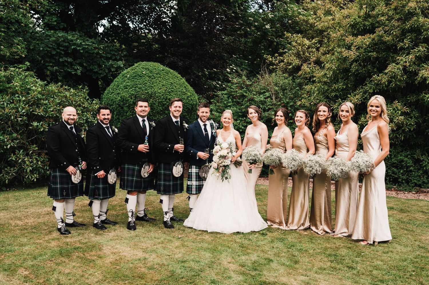 Wedding party poses outdoors with groomsmen wearing traditional Scottish kilts.