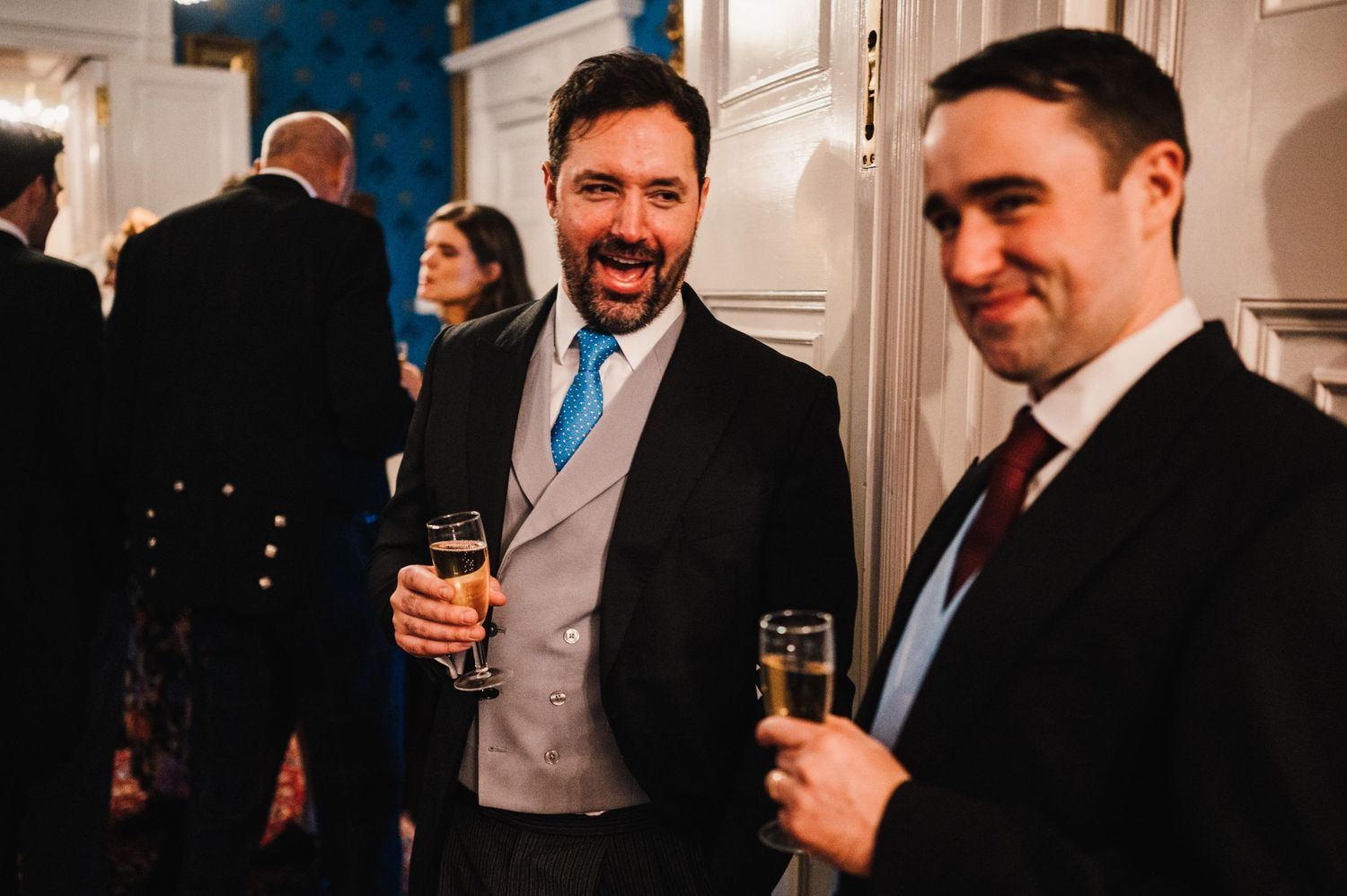 Two well-dressed guests share drinks and laughs during an indoor evening celebration.