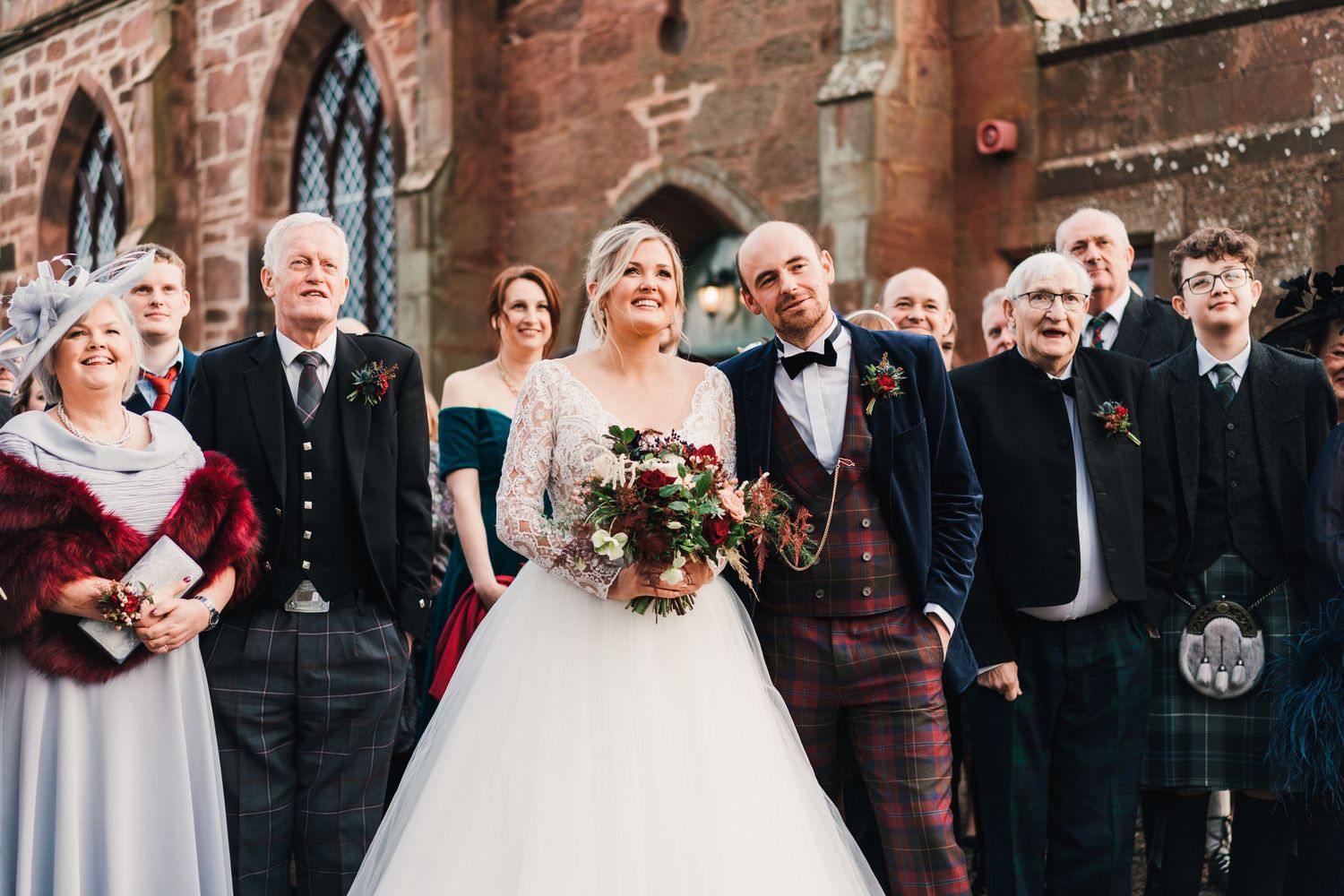 Wedding party in formal attire and kilts poses outside Scottish brick church.