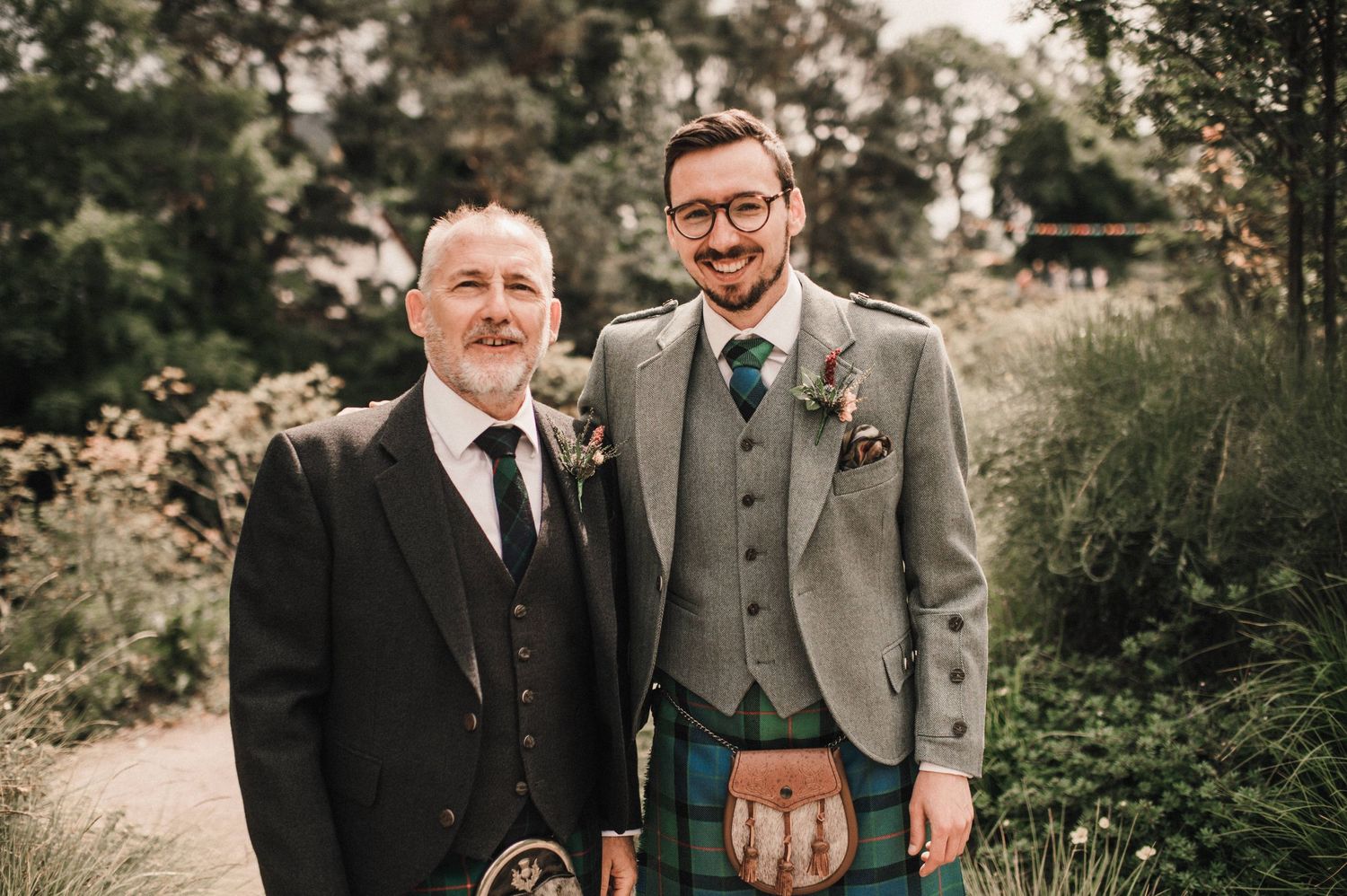 Two people pose together in formal wear with Scottish kilts against a garden backdrop.