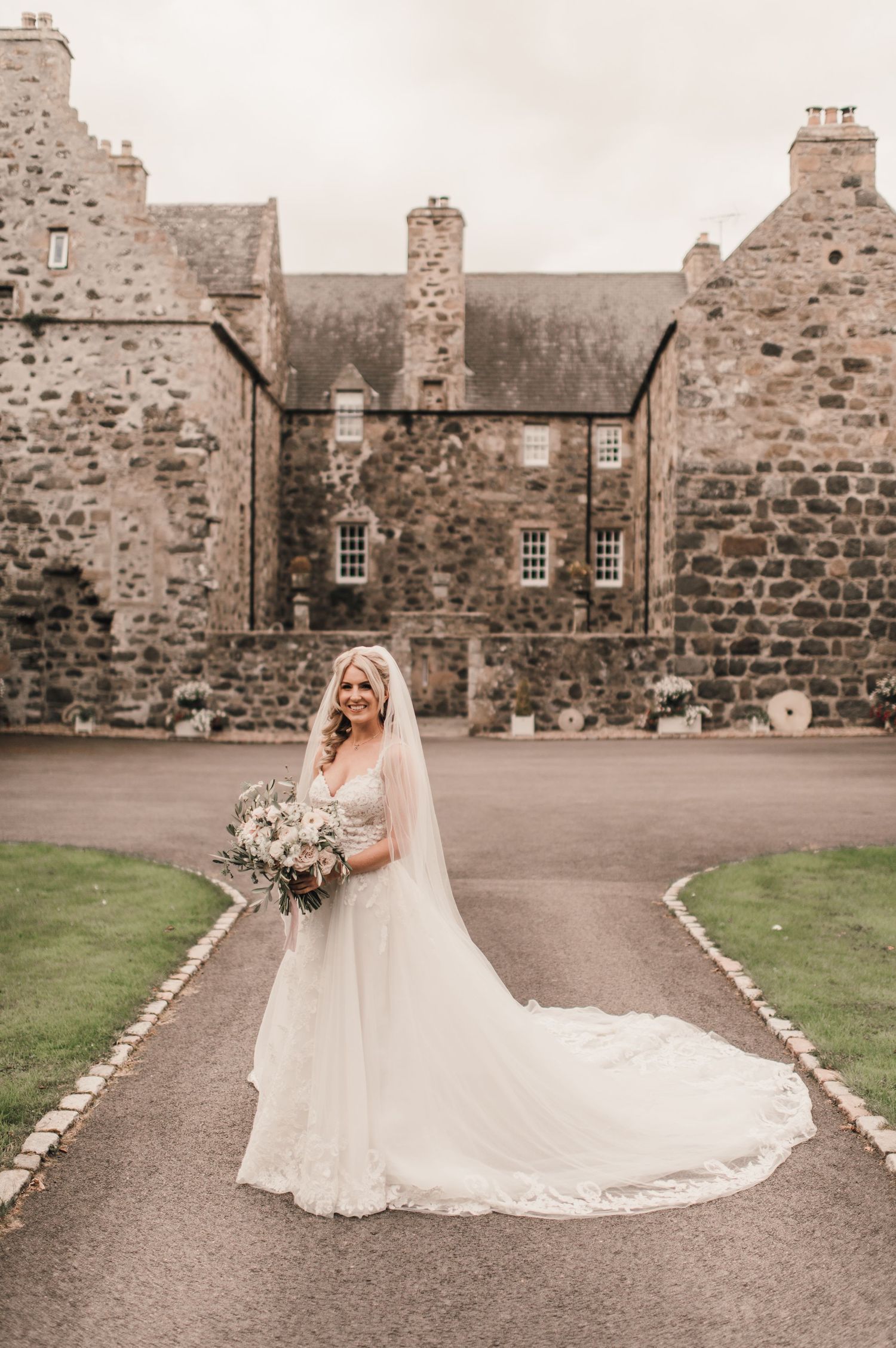 A bride in a flowing white gown poses in front of a majestic Scottish stone castle.