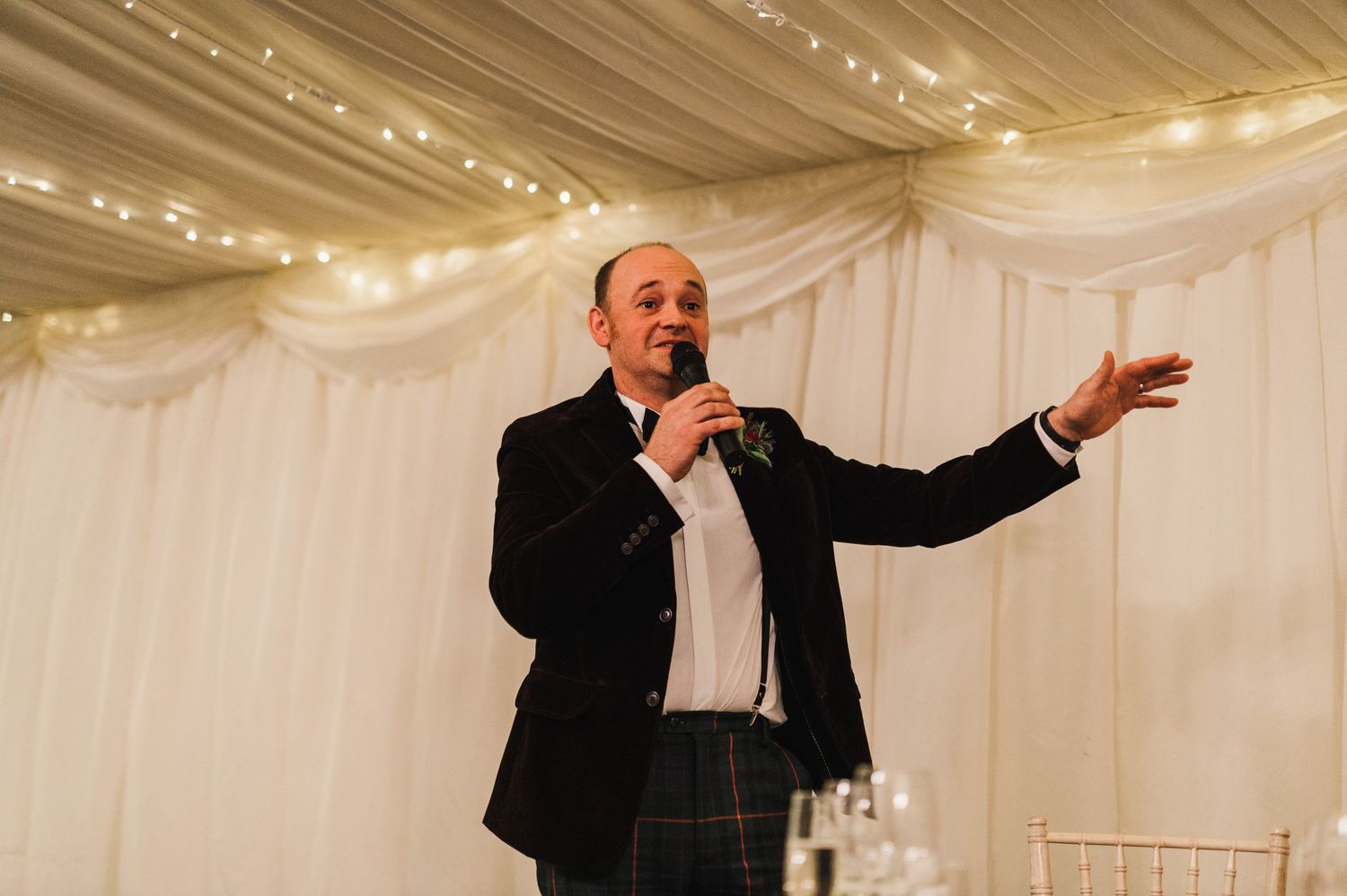 A speaker gestures enthusiastically during a wedding reception speech in a decorated tent venue.