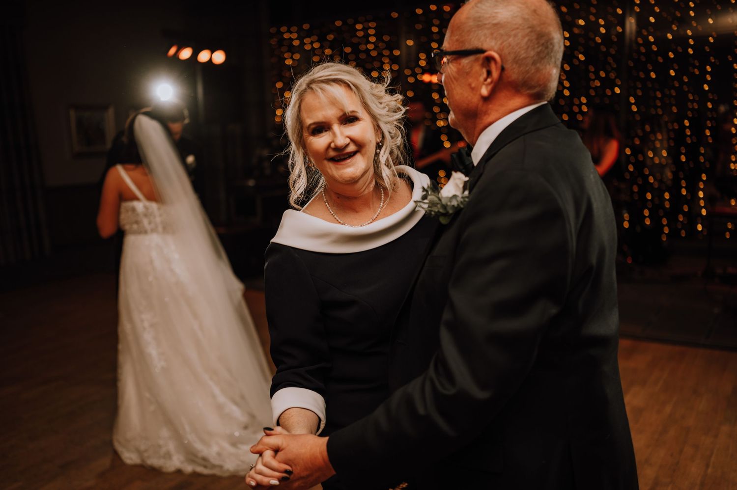 Guests dance and smile on wedding reception dance floor with twinkle lights in background.