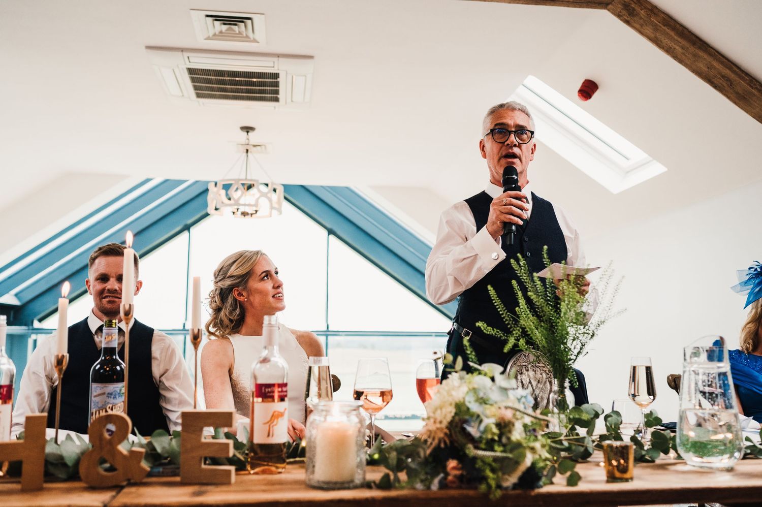 Wedding reception scene in a bright venue with table decorations and greenery.
