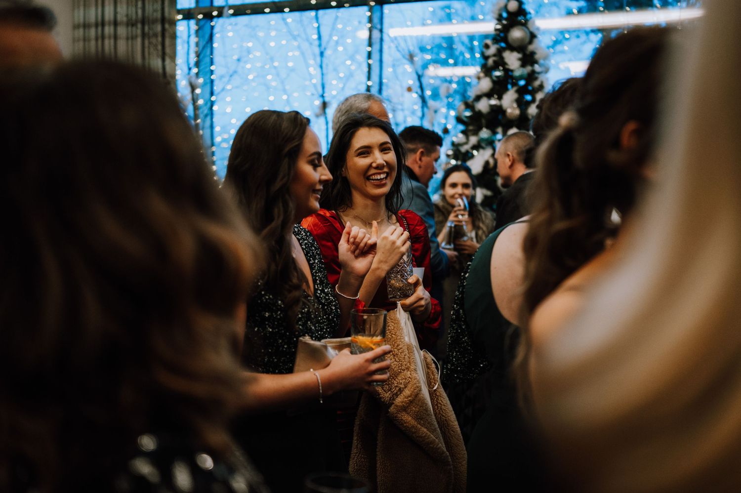 Party guests mingle and celebrate with drinks under blue decorative lighting and a Christmas tree.