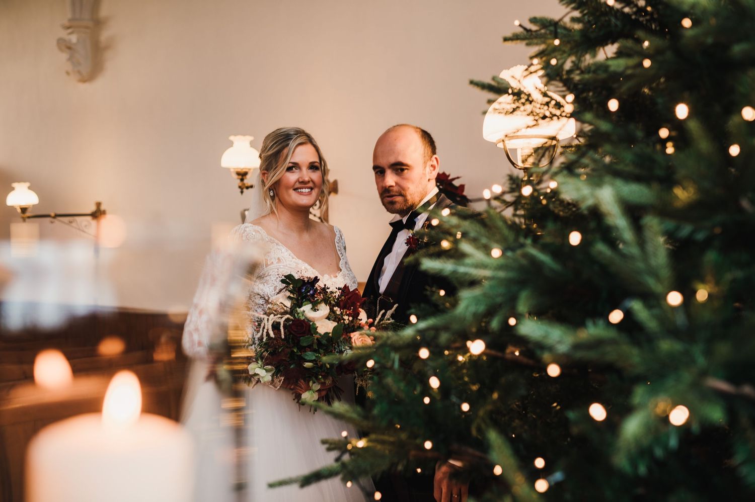 Couple shares intimate moment by candlelit Christmas tree during winter wedding celebration.