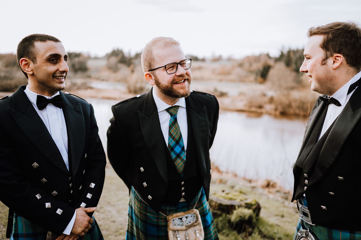 Three people in traditional Scottish formal wear talking by a lake on an overcast day.