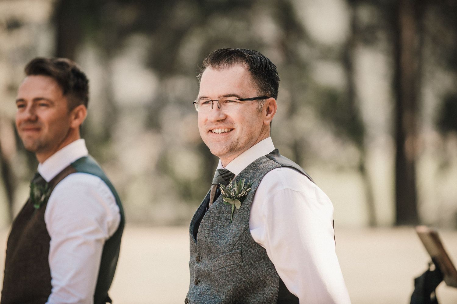 Two groomsmen in tweed vests smile during an outdoor wedding celebration.