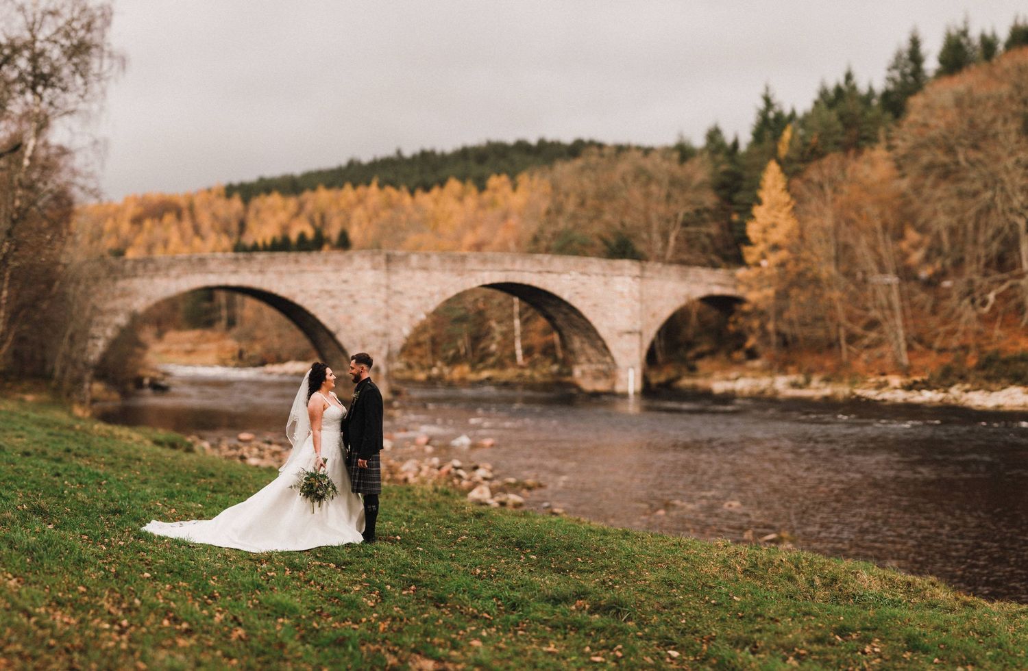 Wedding couple stands on grassy riverbank with historic stone bridge in background.