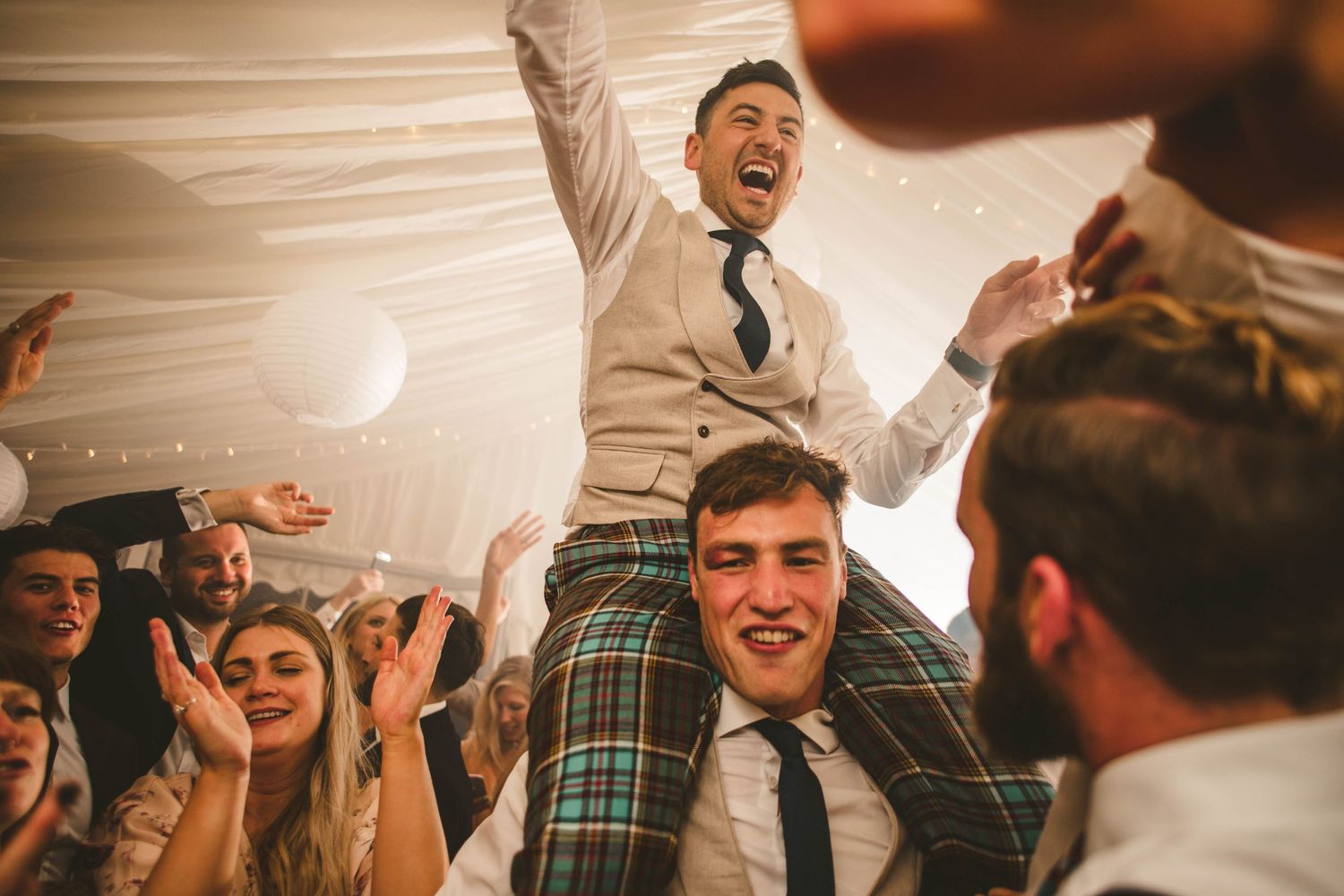Energetic celebration with partygoers dancing and cheering at a wedding reception under draped fabric ceiling.
