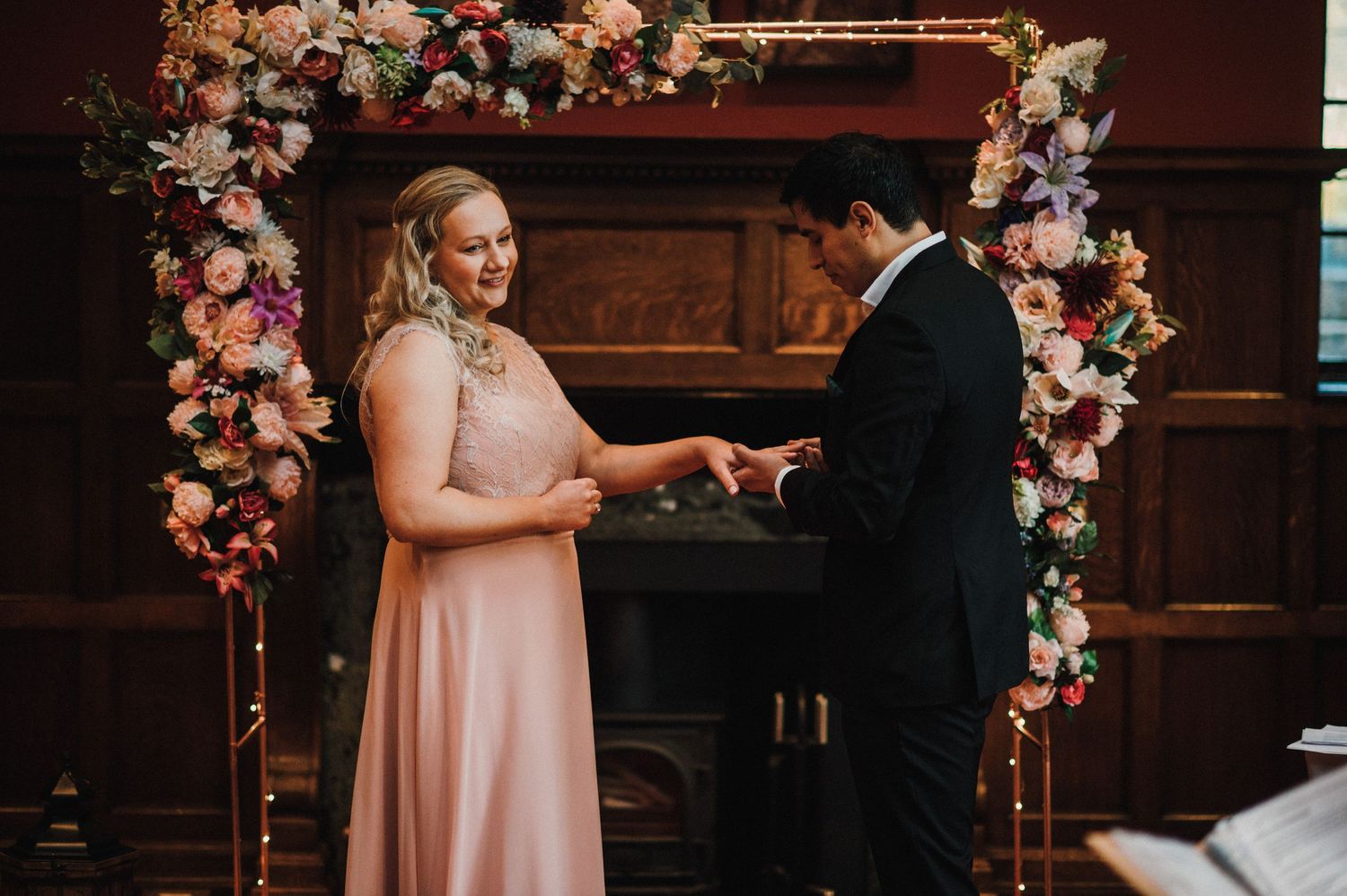 A romantic exchange of rings under a floral arch during an indoor wedding ceremony.