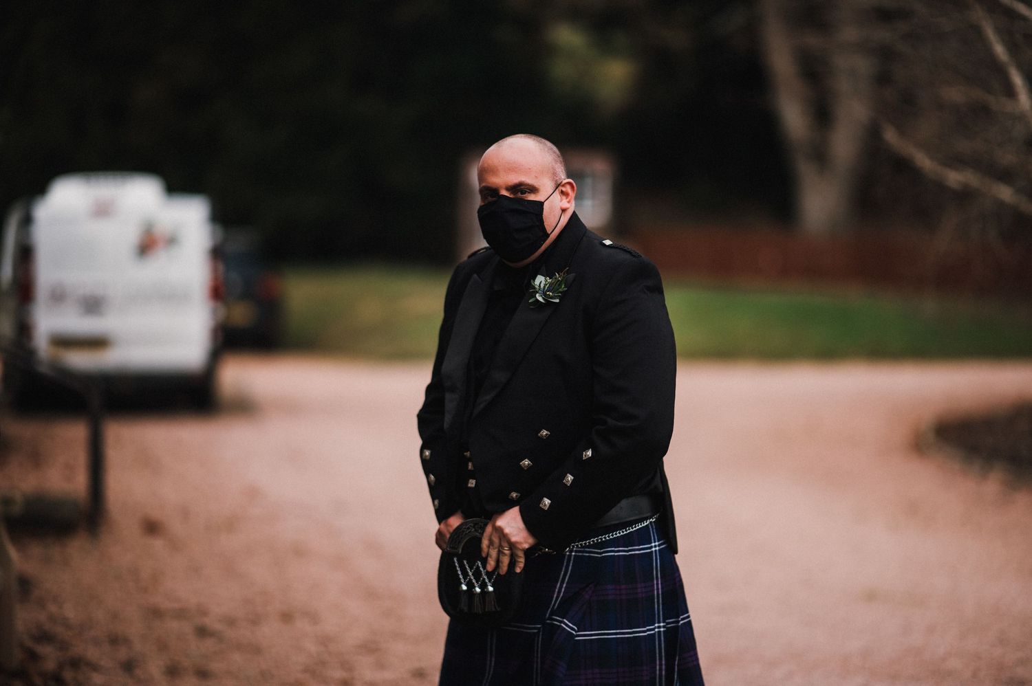Person in traditional Scottish kilt and black coat wearing face mask outdoors.
