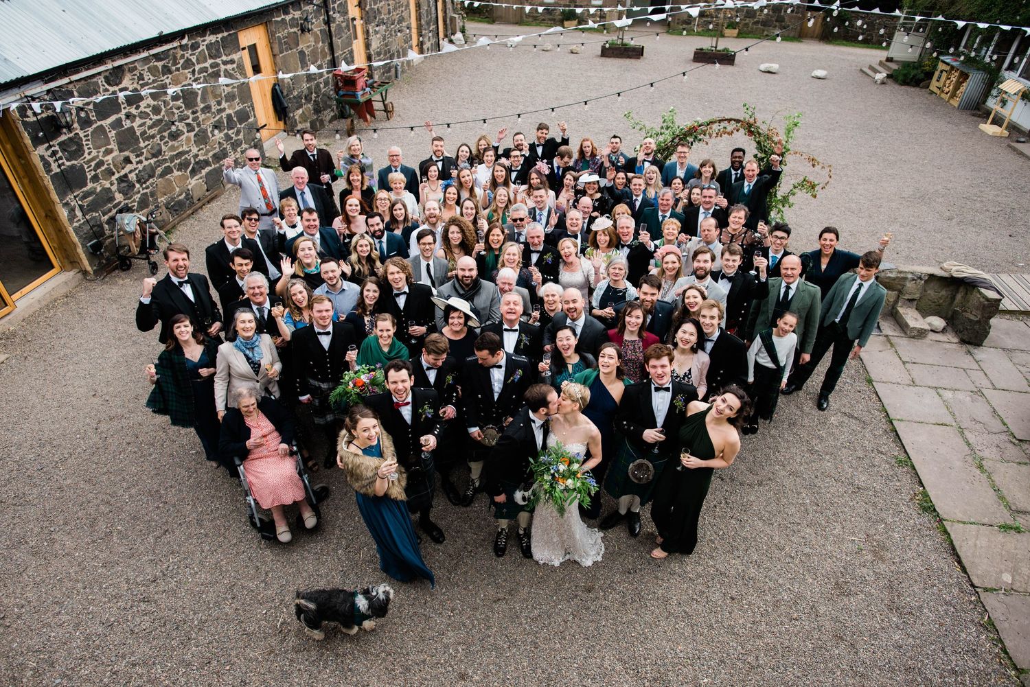 Aerial group photo of wedding party and guests in traditional Scottish formal wear at stone venue.