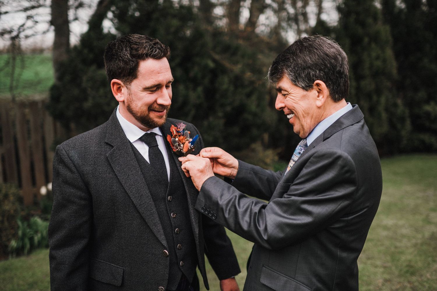 Two men in formal suits share a special moment adjusting a boutonniere at a Scottish wedding ceremony.