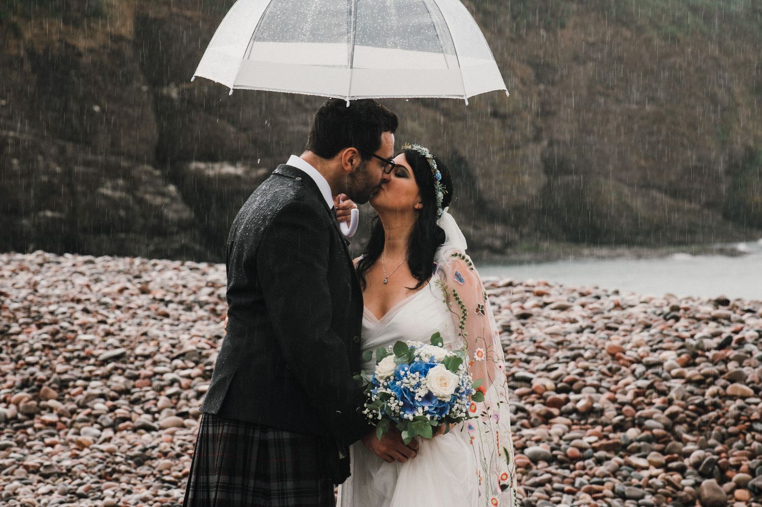 Romantic wedding couple sharing an intimate moment under a clear umbrella on a rocky coastline.
