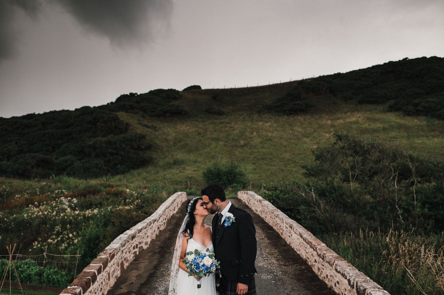 A wedding couple shares an intimate moment on a rustic stone bridge with hills in background.