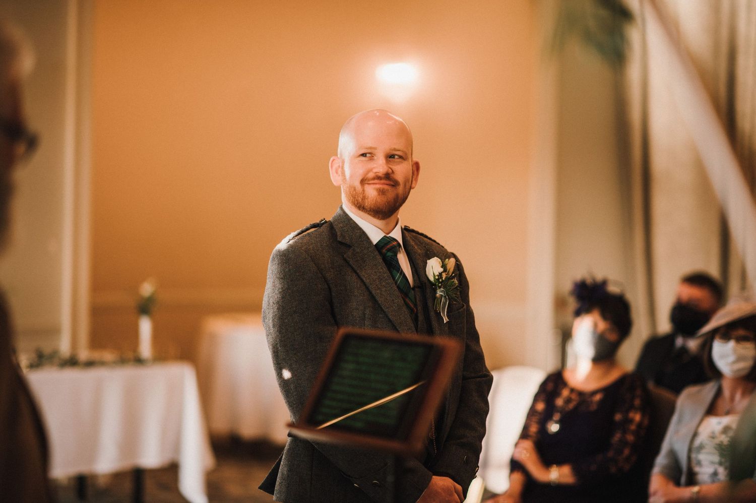 Wedding guest wearing traditional Scottish kilt stands smiling in an elegant reception venue.