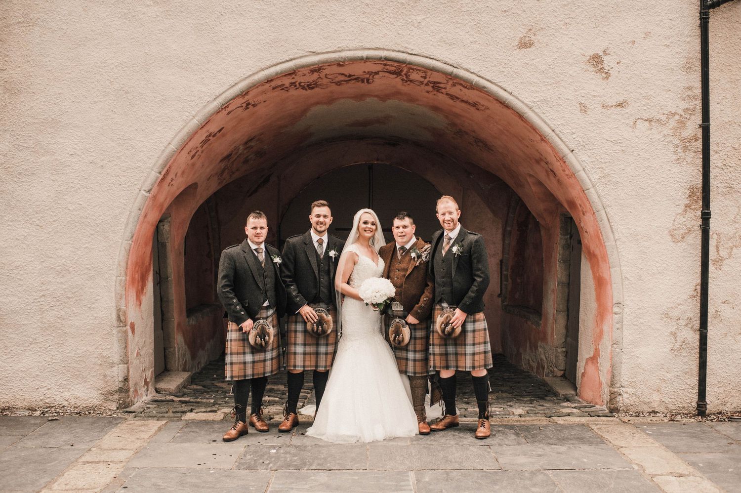 Wedding party in Scottish formal wear poses under stone archway with classic architectural details.