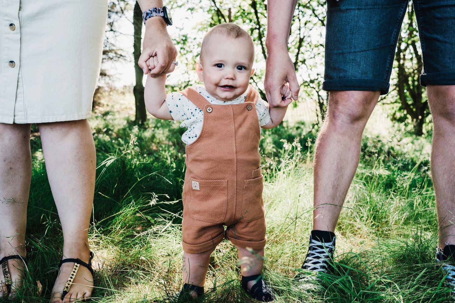 A toddler wearing brown overalls stands supported between two adults in a sunny outdoor setting with grass and greenery.