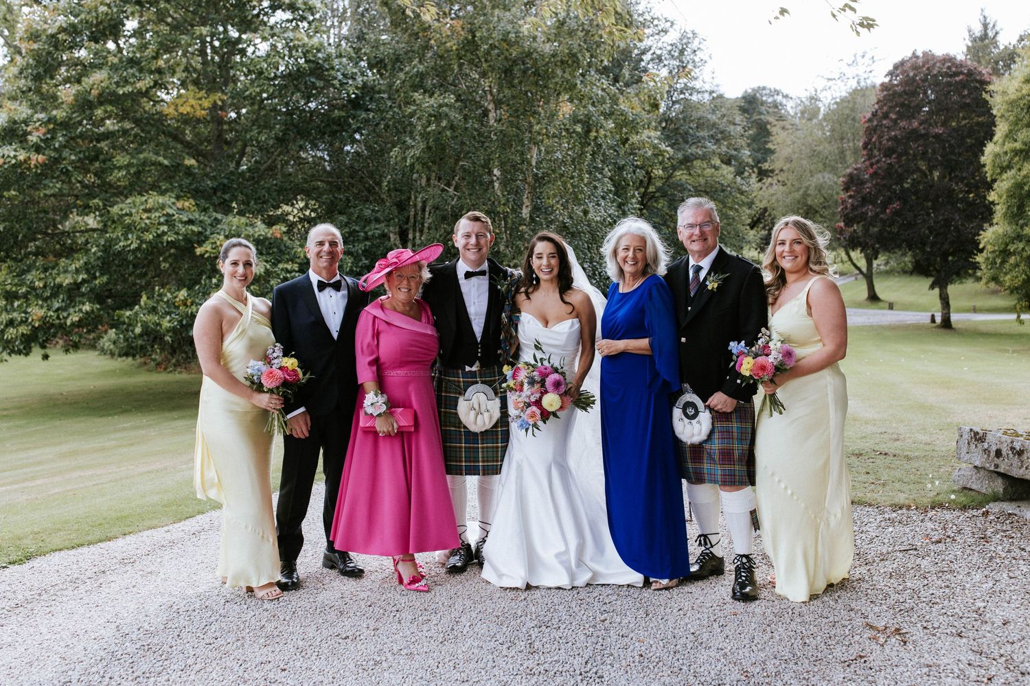 Wedding party in traditional Scottish attire poses on gravel path with lush green garden backdrop.