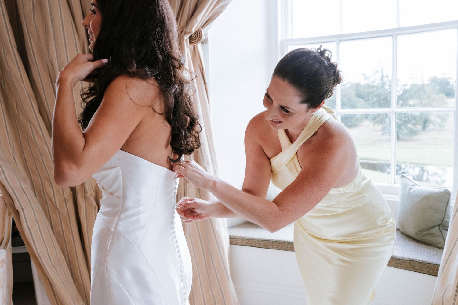 Final adjustments being made to a white wedding dress in front of a window.