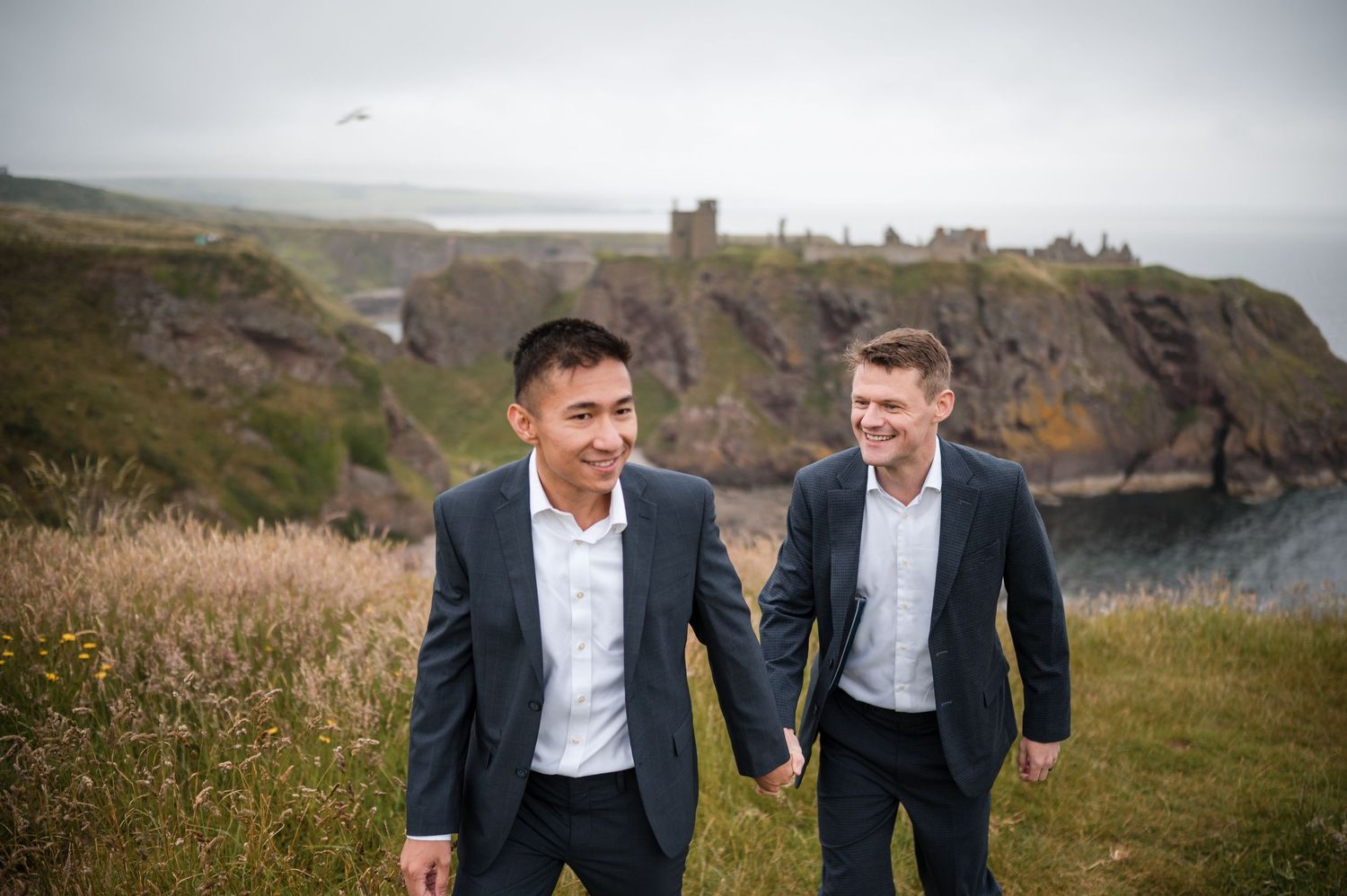 Same Sex Wedding PhotographA couple shares a playful moment on a scenic coastal hillside with historic castle ruins visible in the distance.