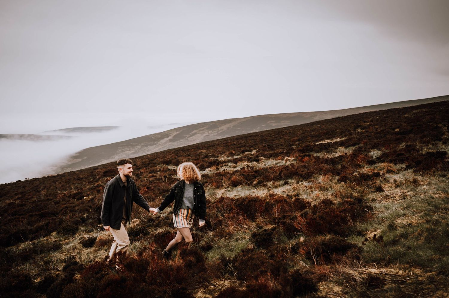 Engagement photography session Aberdeen. A couple stands together on a rugged hillside overlooking misty mountains.