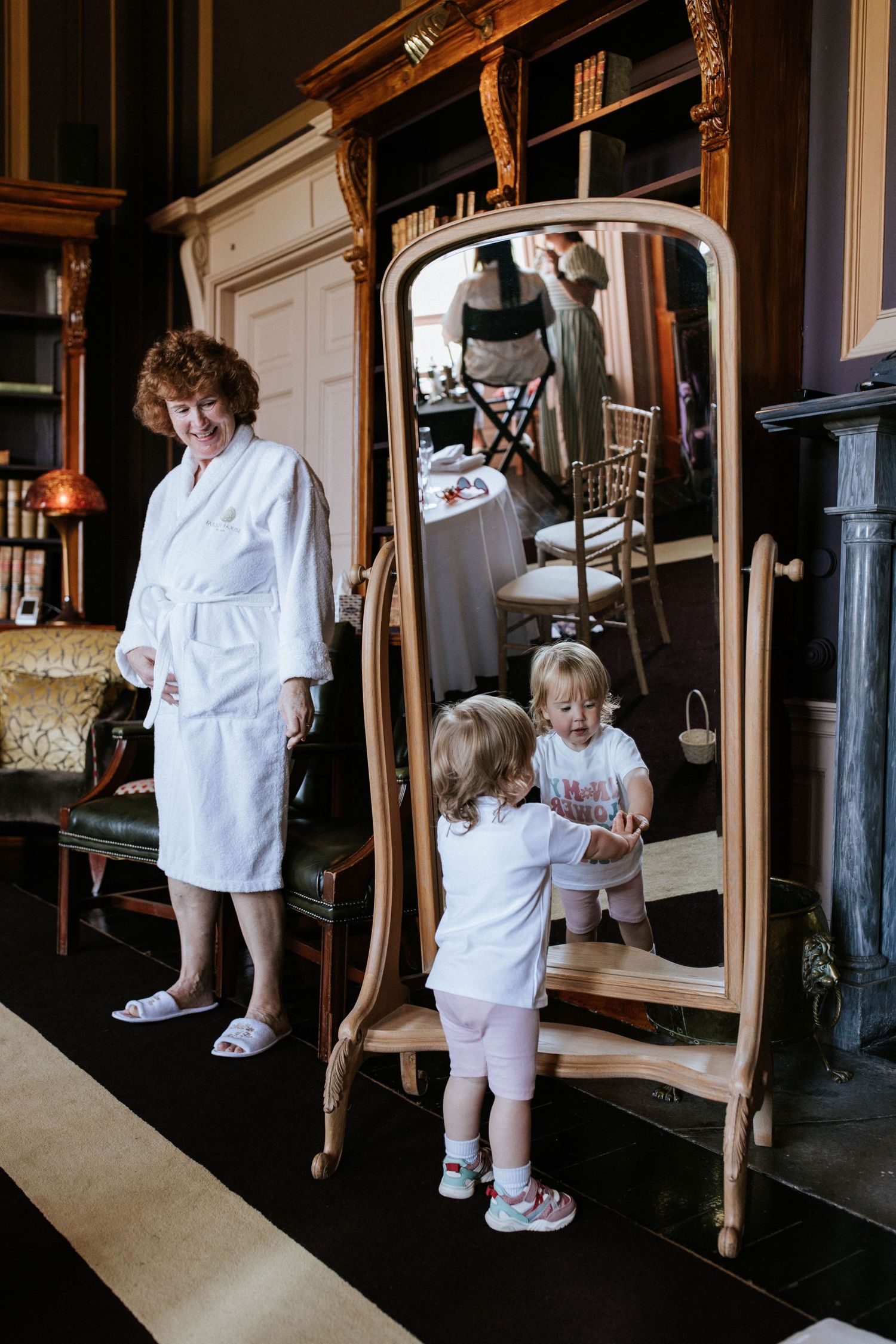 People in white robes standing by an ornate full-length mirror in a vintage-styled room.