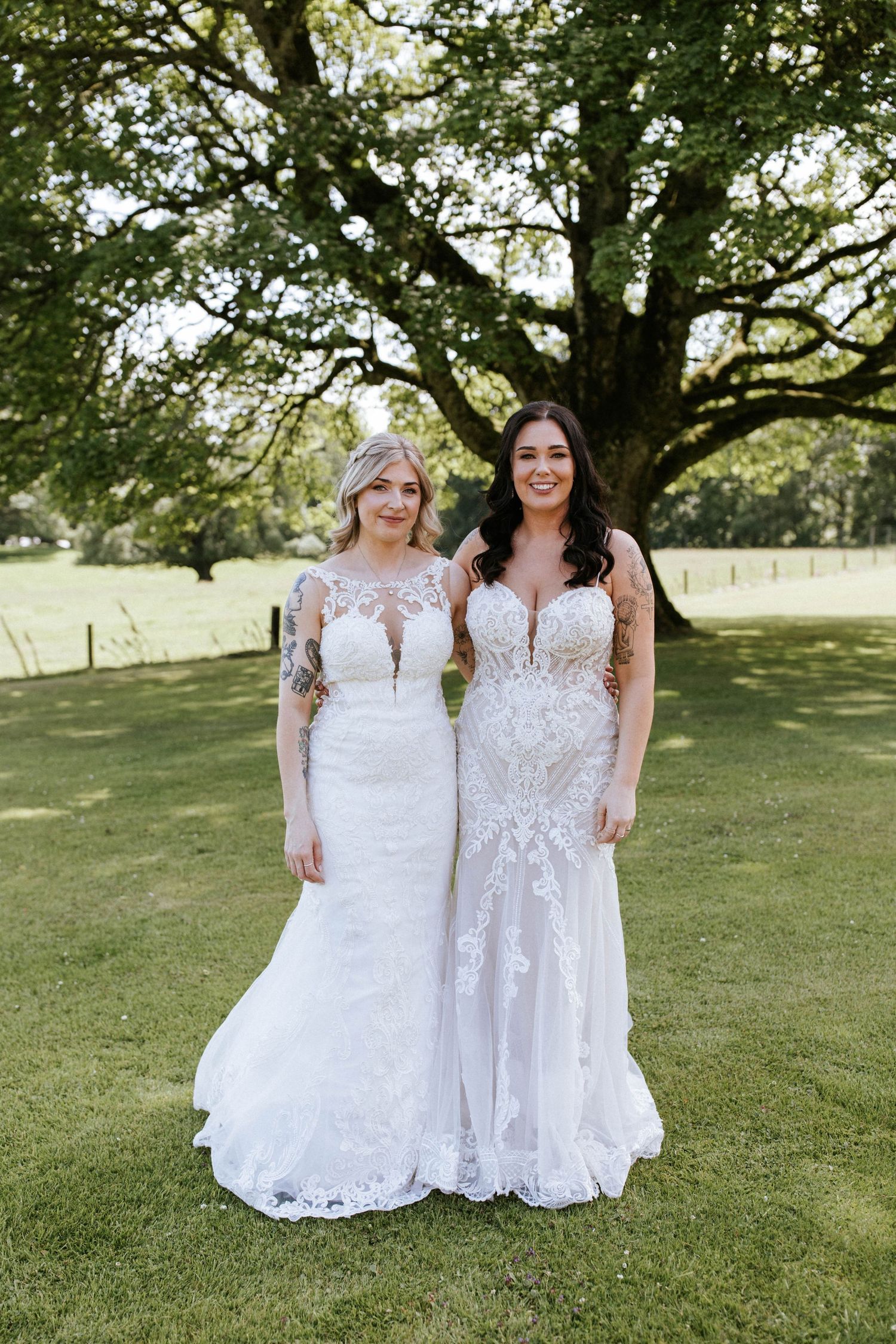 Two people in elegant white lace wedding dresses pose together on a grassy lawn under a large tree.