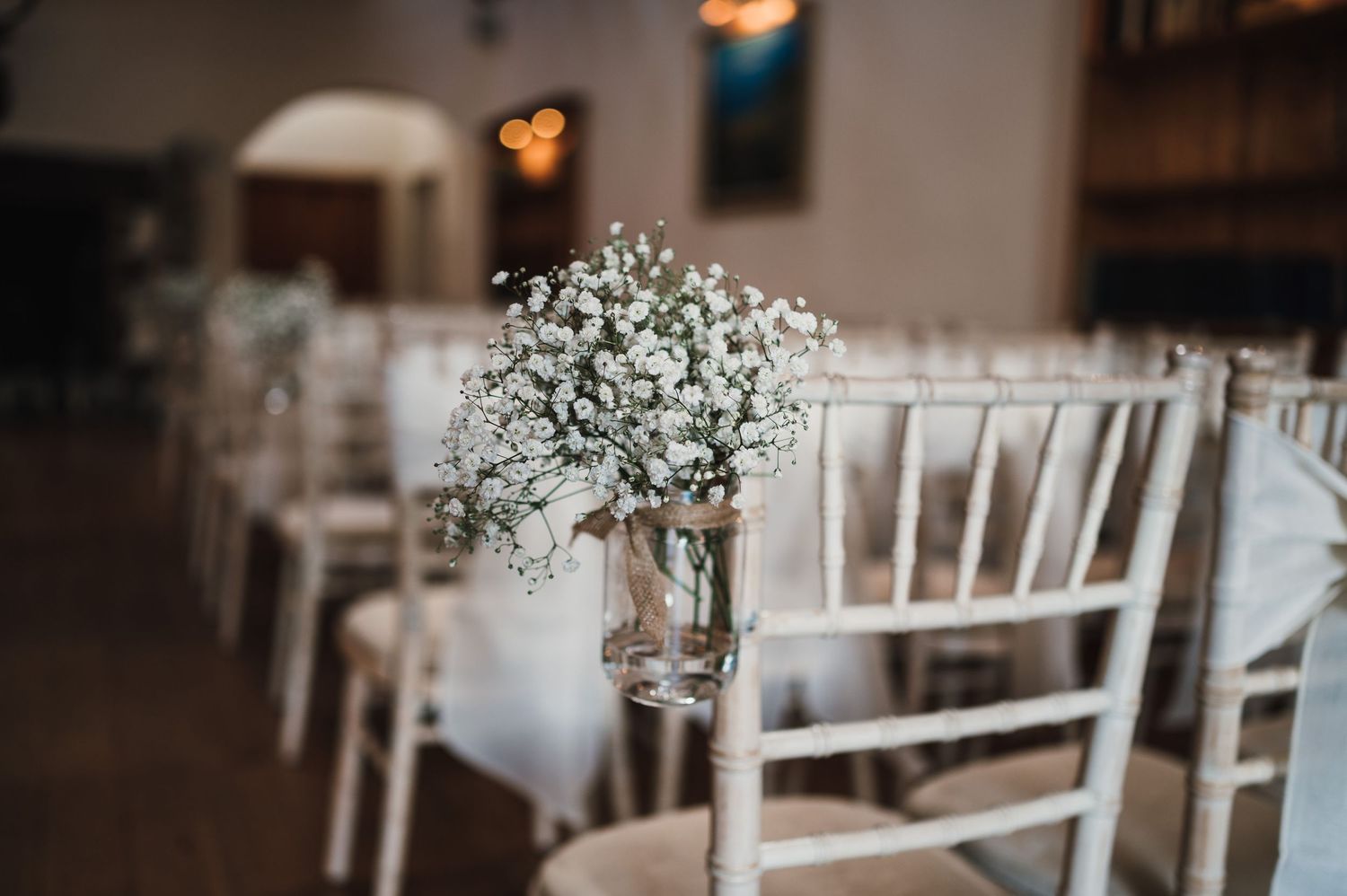 White baby's breath floral centerpieces in glass vases adorn white Chiavari chairs at a wedding venue.