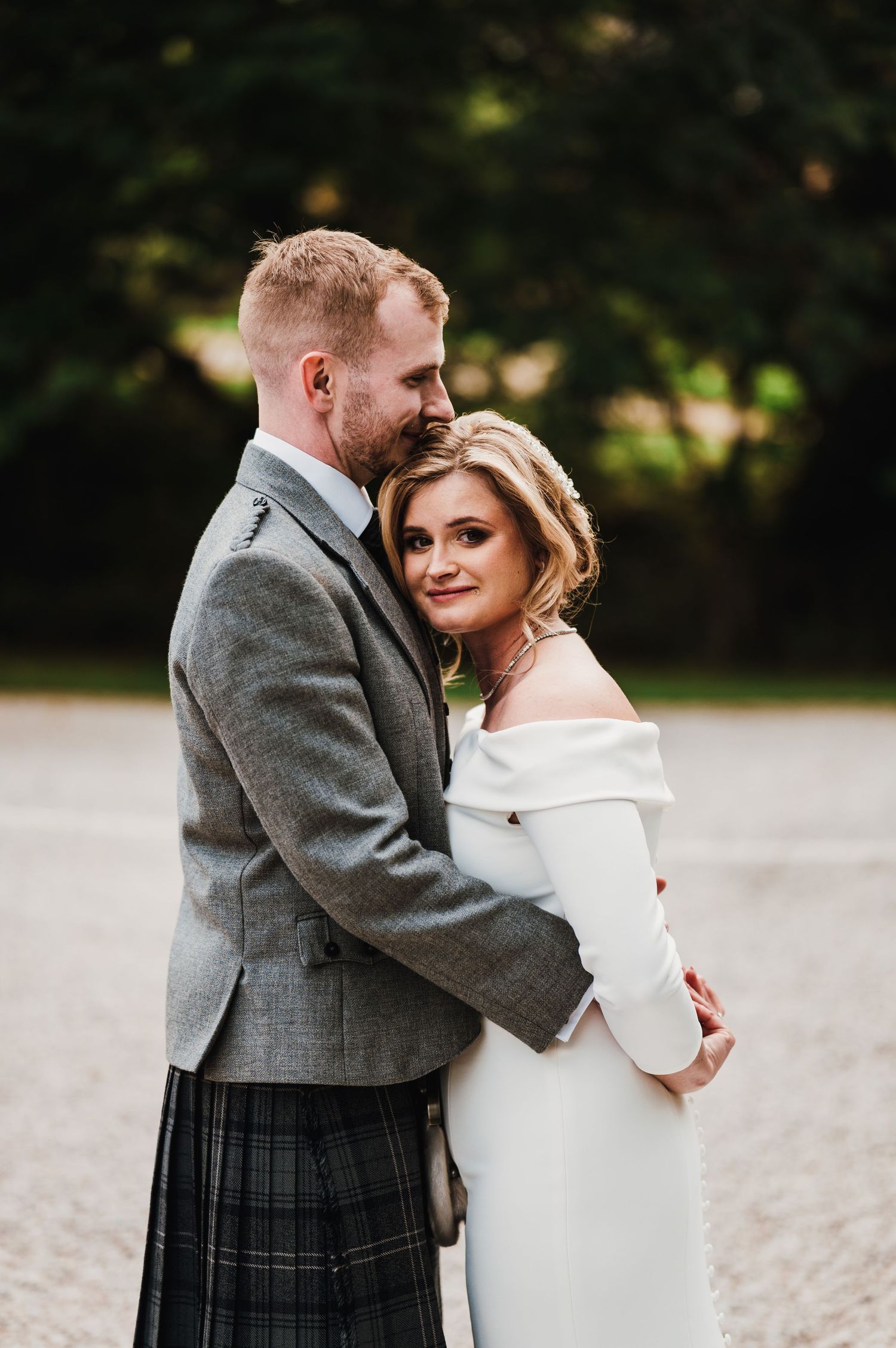 A couple embraces outdoors, with the person wearing a traditional Scottish kilt and gray jacket.