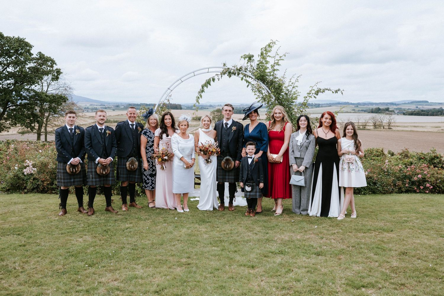 Wedding party and guests pose for formal group photos on a grassy hillside with scenic views.