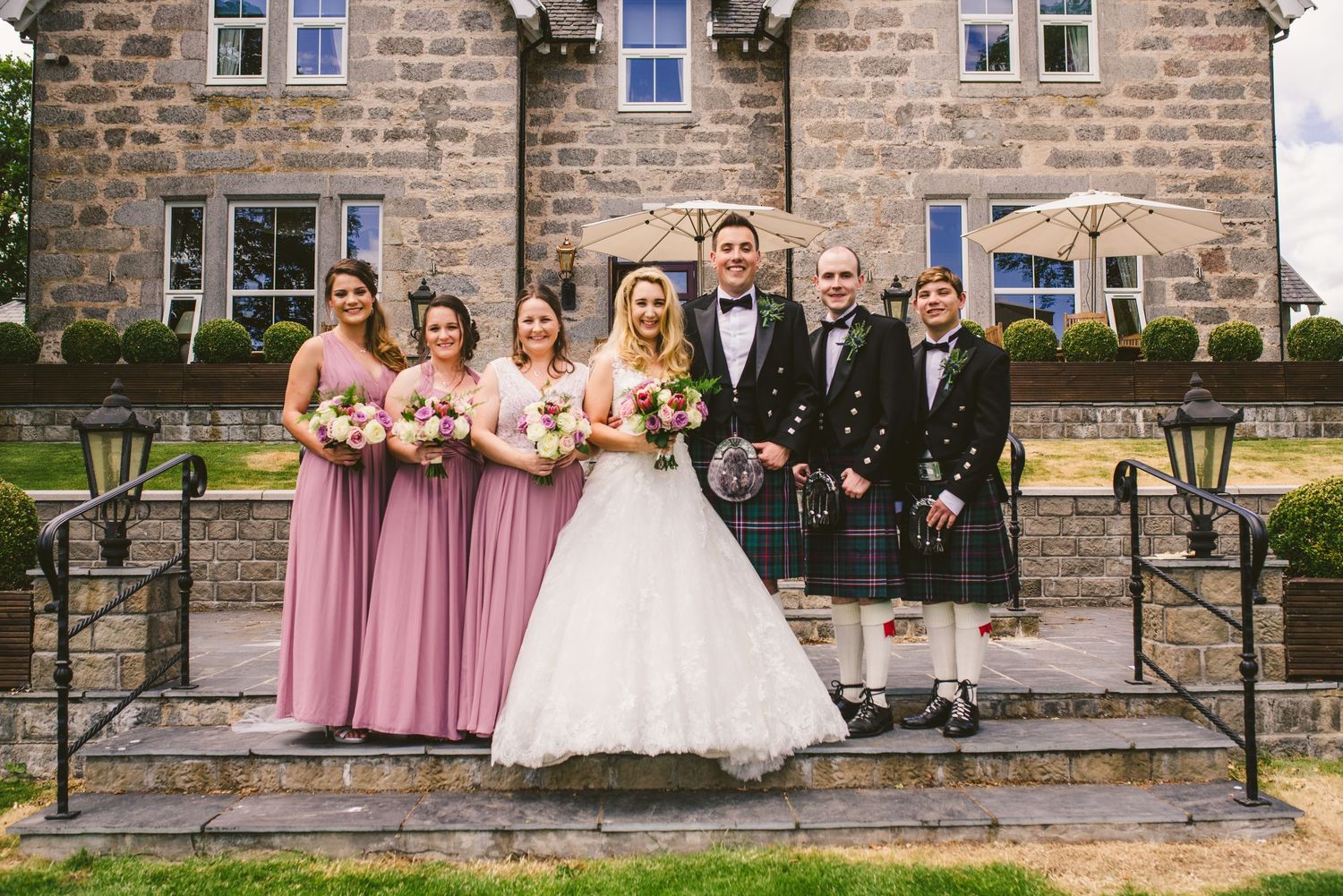 Wedding party poses outside historic stone building with manicured grounds.