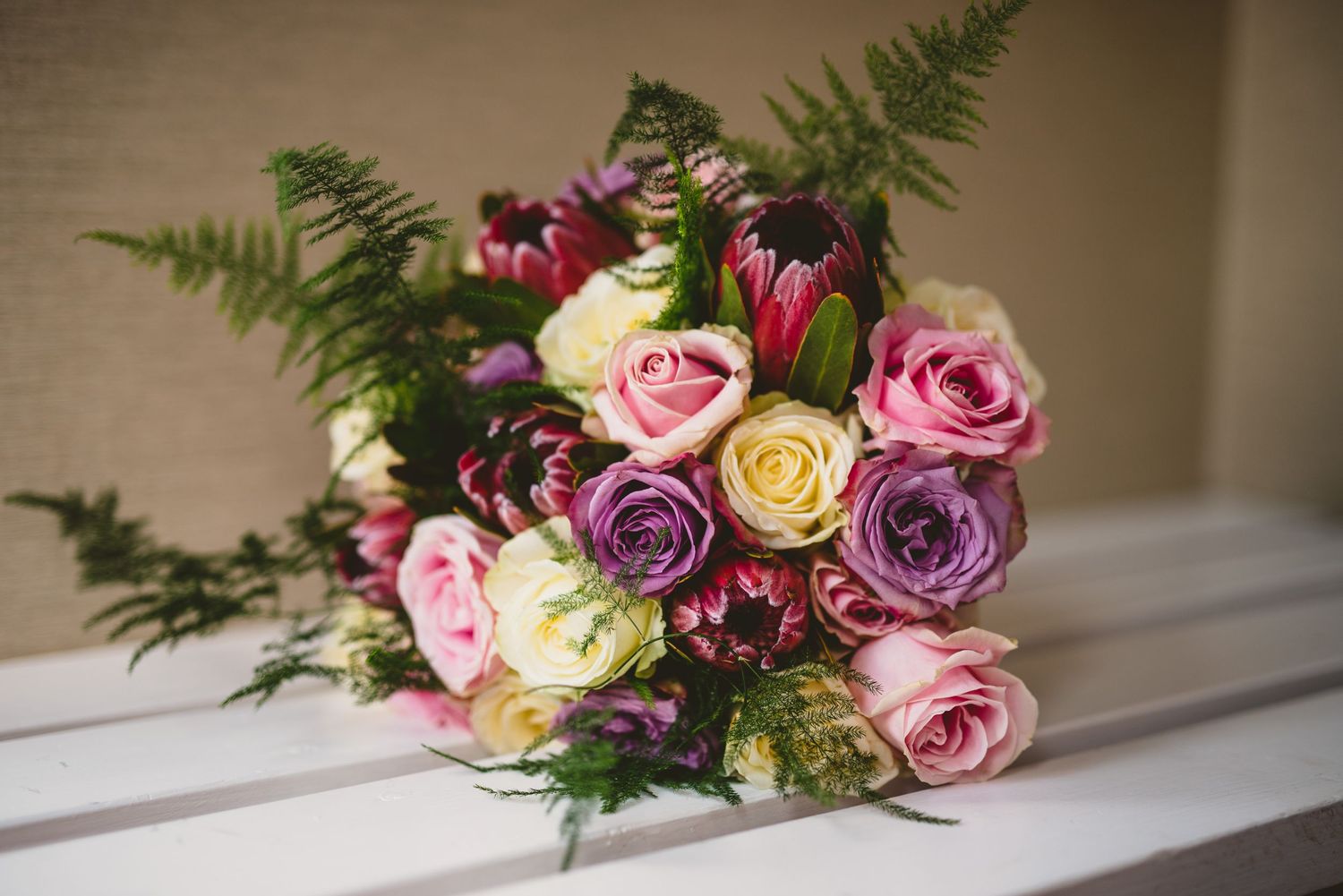 Colorful bouquet of roses and ferns arranged on white ledge with soft natural lighting.