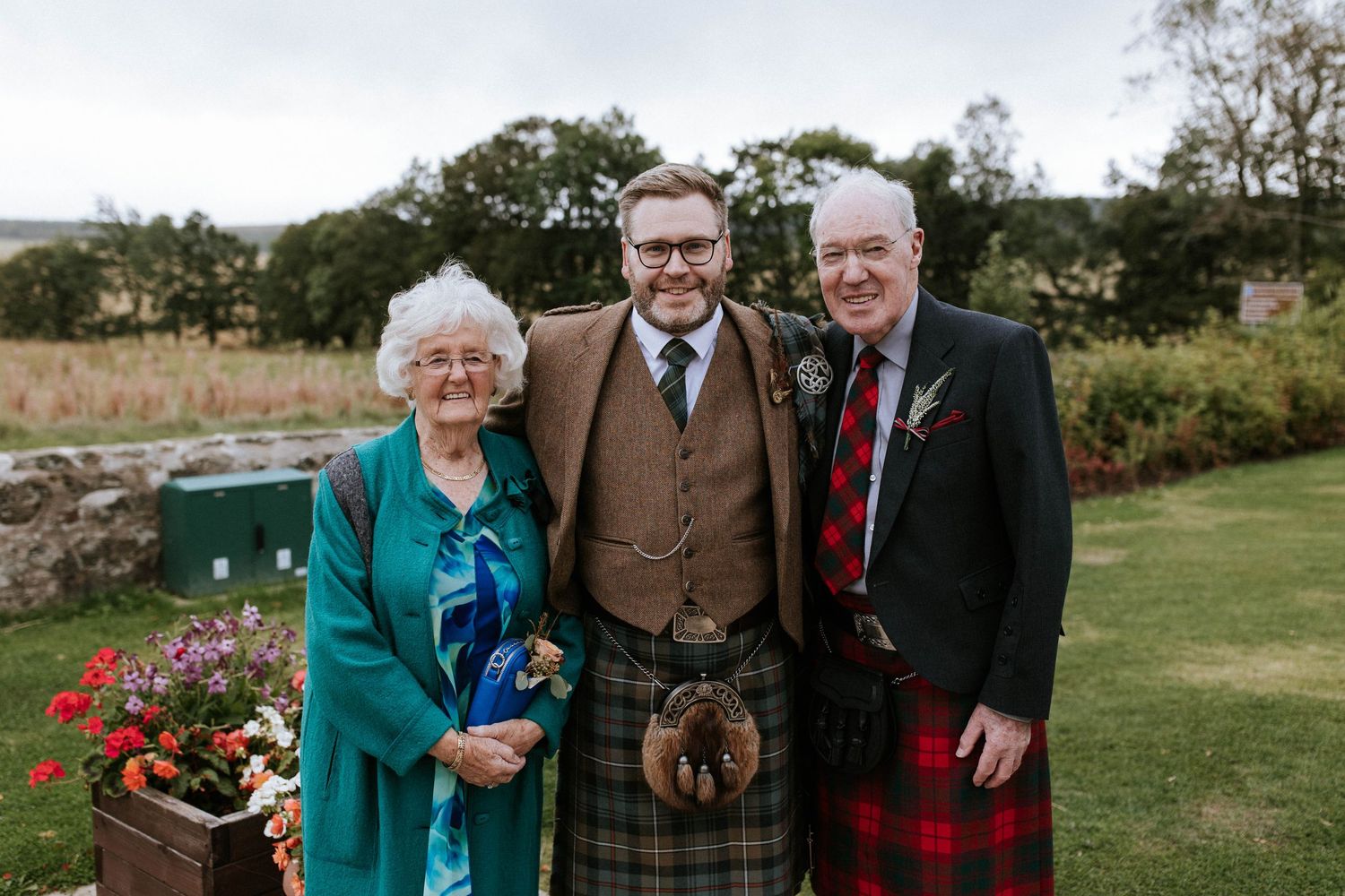 Three generations gather outdoors in traditional Scottish attire with tartan patterns and tweed jackets.