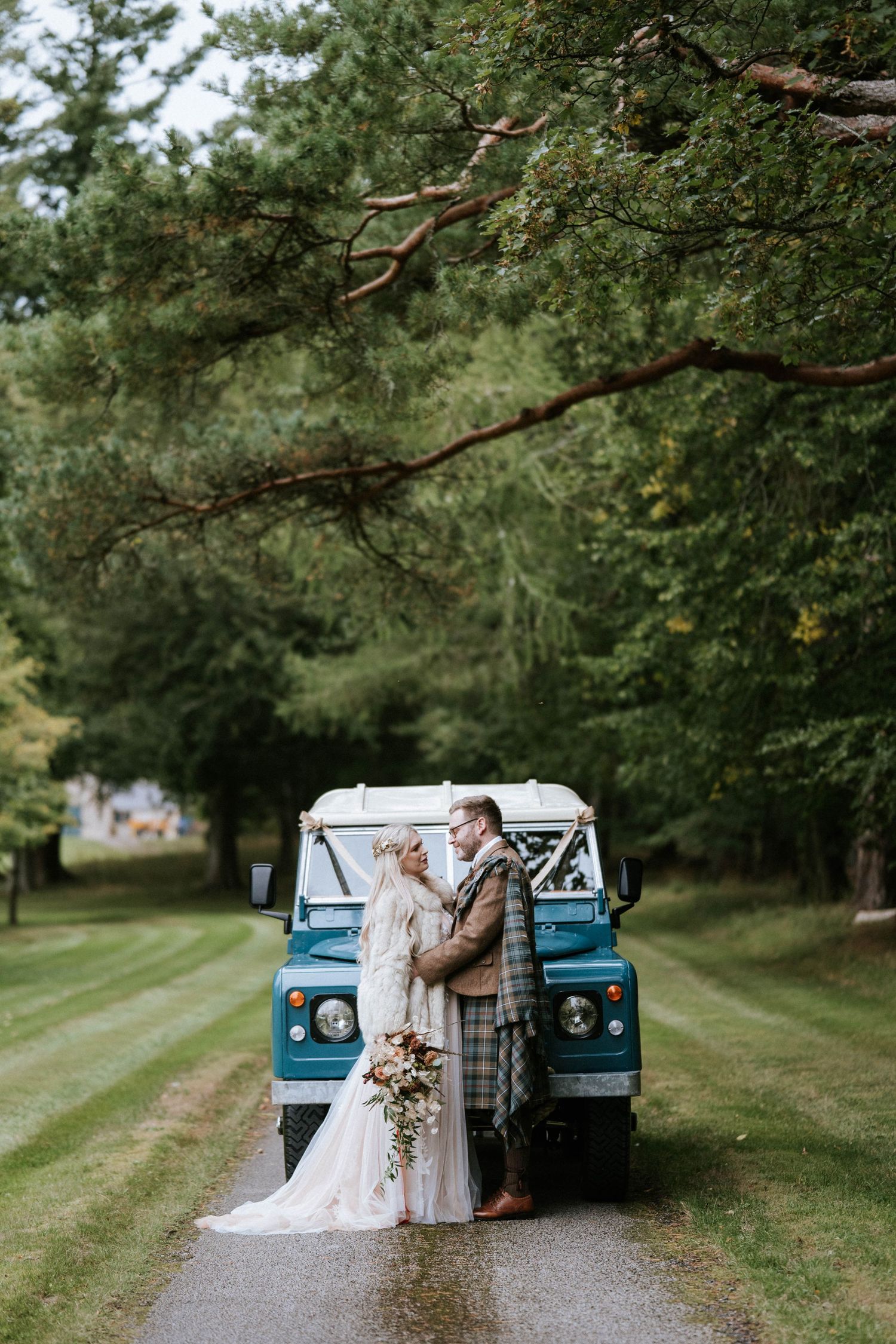A vintage blue pickup truck serves as a romantic backdrop for a rustic outdoor wedding photoshoot in a wooded setting.