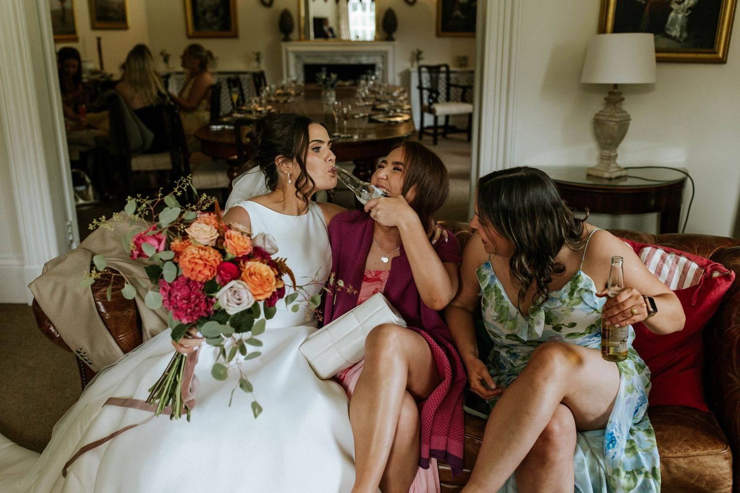 Wedding party relaxes on a couch with colorful bouquets and laughter.