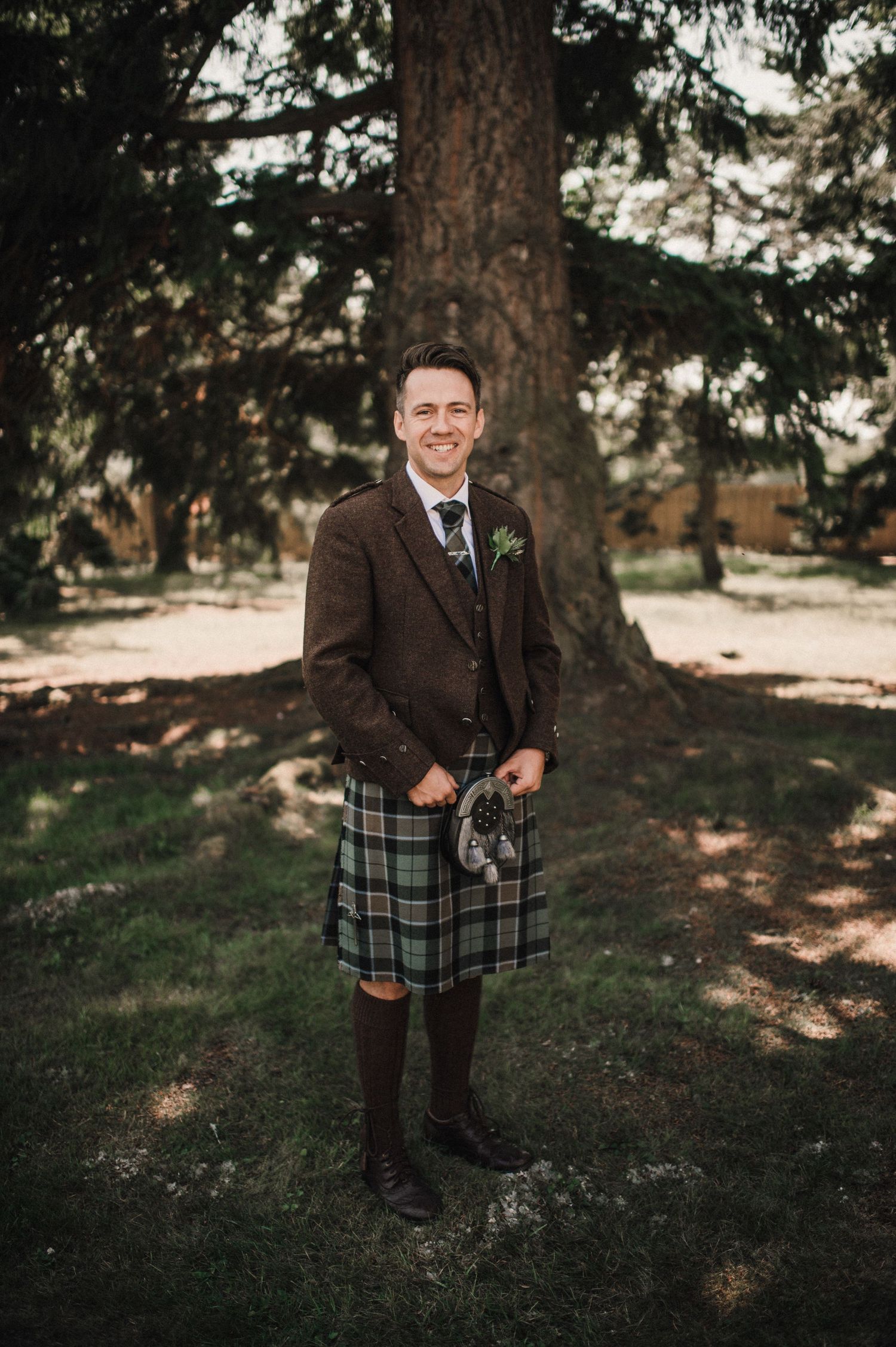 Person in brown tweed jacket and traditional kilt poses beside large tree trunk.