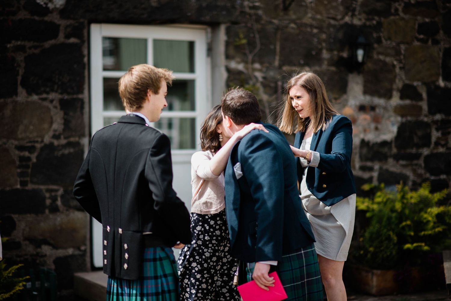 Party guests mingle outside a stone building with traditional Scottish kilts and formal attire.