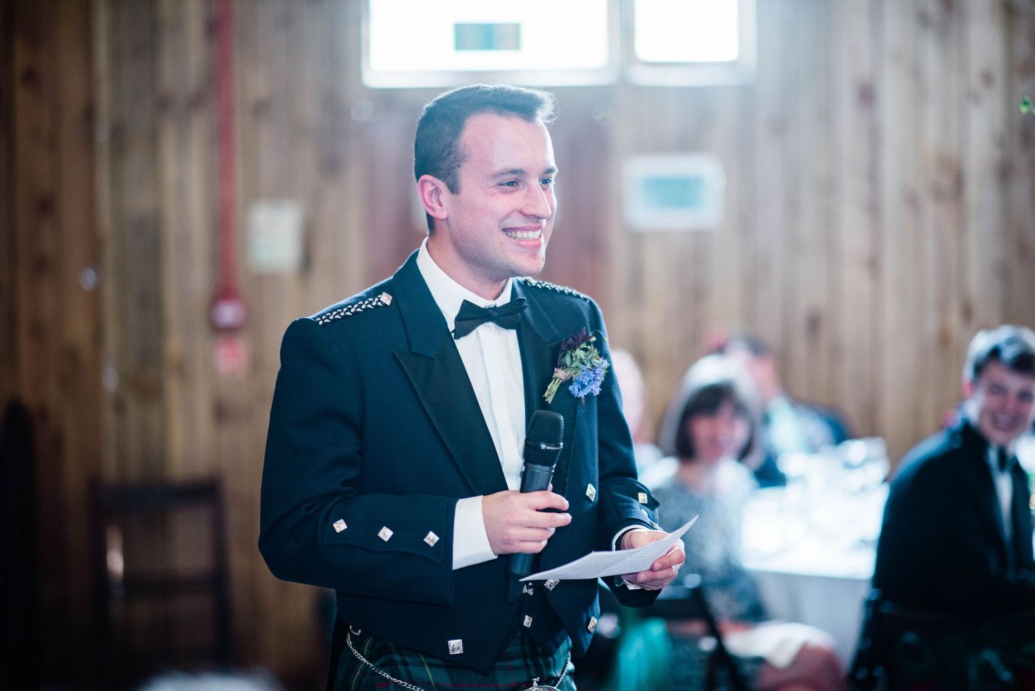 Guest in formal Scottish attire gives toast during indoor wedding reception.