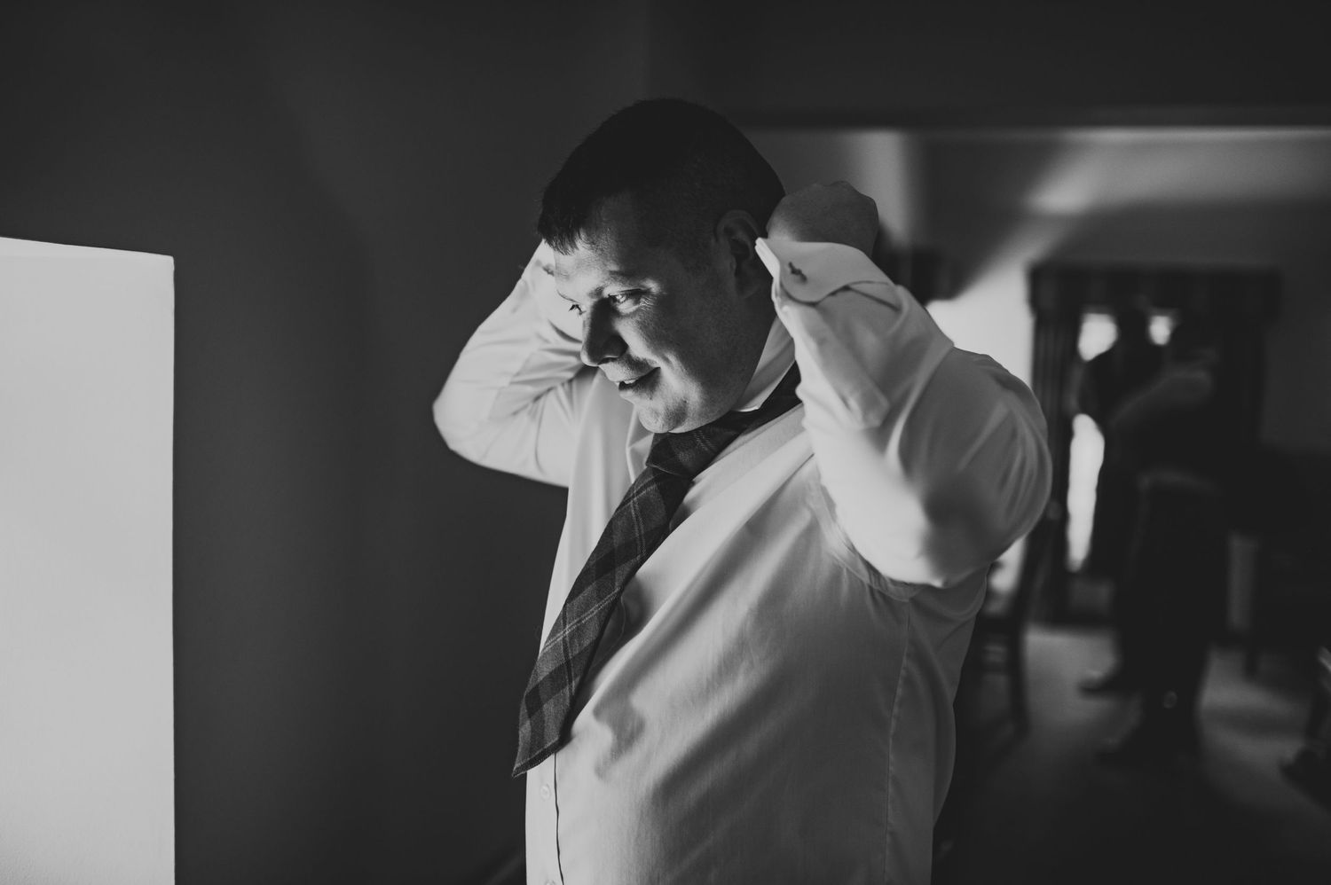 A man in a white dress shirt adjusts his collar in a dimly lit room.