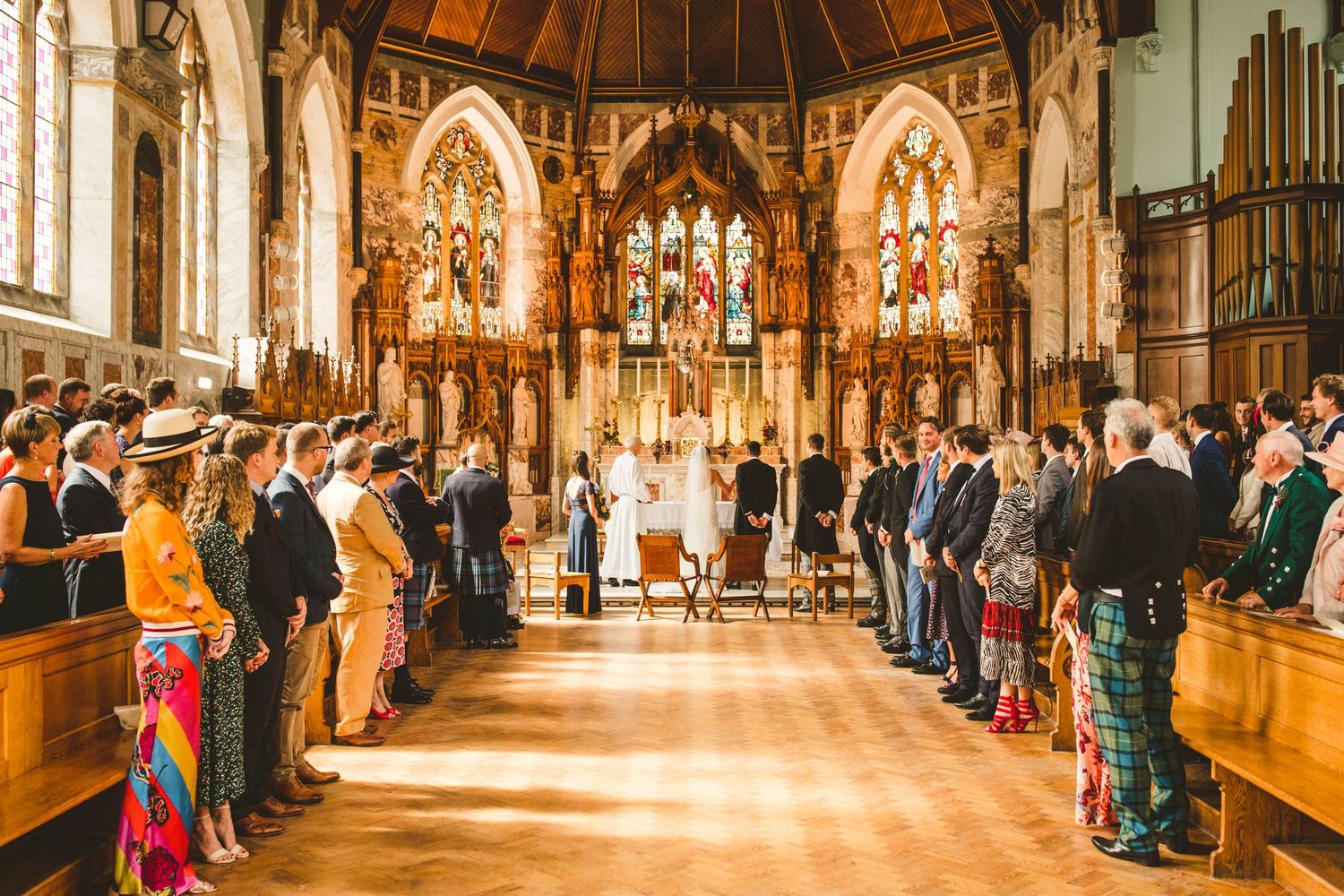 Wedding guests stand in rows inside an ornate church with stained glass windows and gothic architecture.