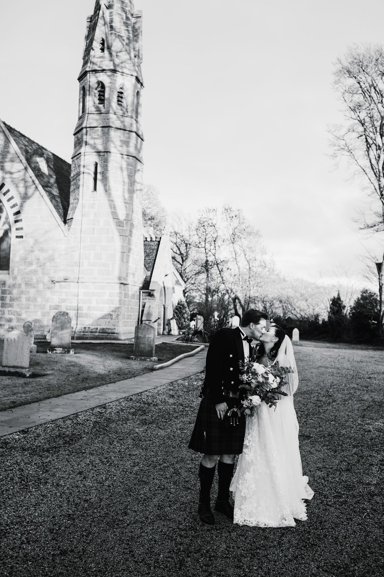 A wedding couple poses in front of a historic stone church in traditional Scottish and bridal attire.