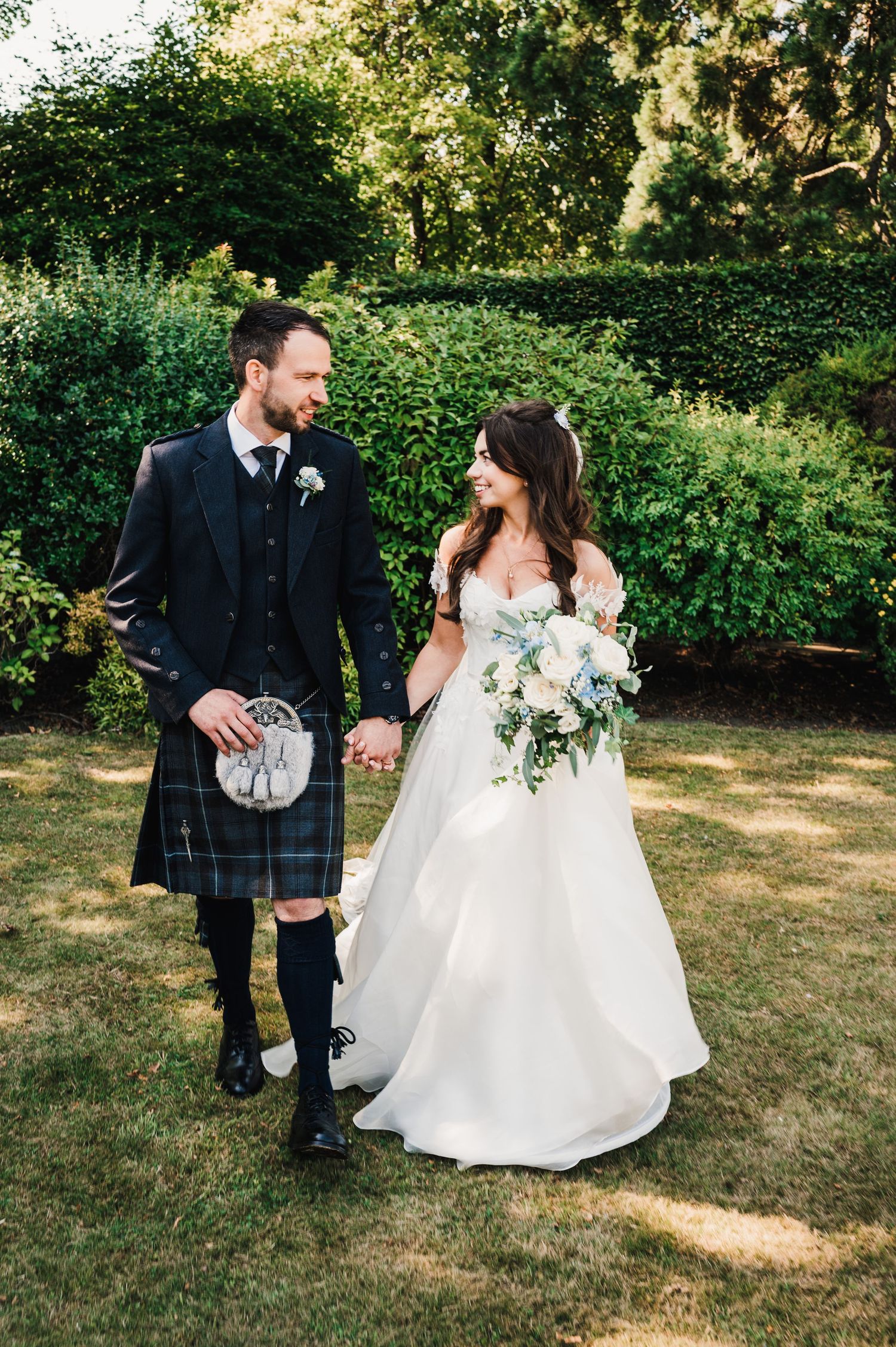 Scottish-themed wedding couple walking hand in hand with kilt and white gown in garden.