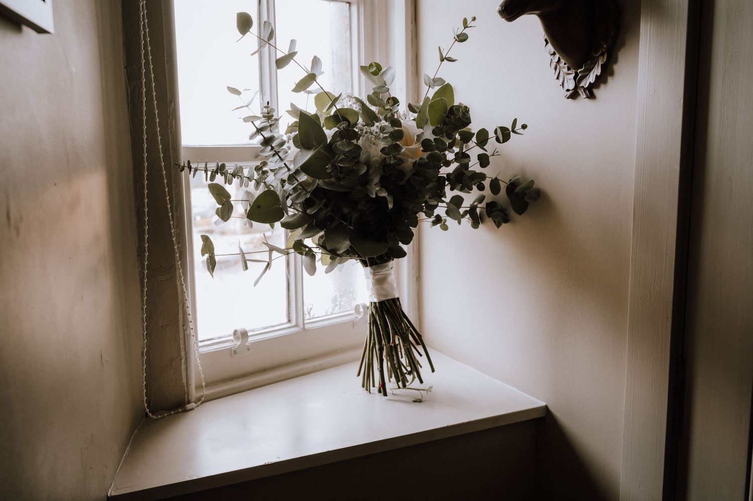 An elegant white floral bouquet with eucalyptus sits on a windowsill in moody lighting.