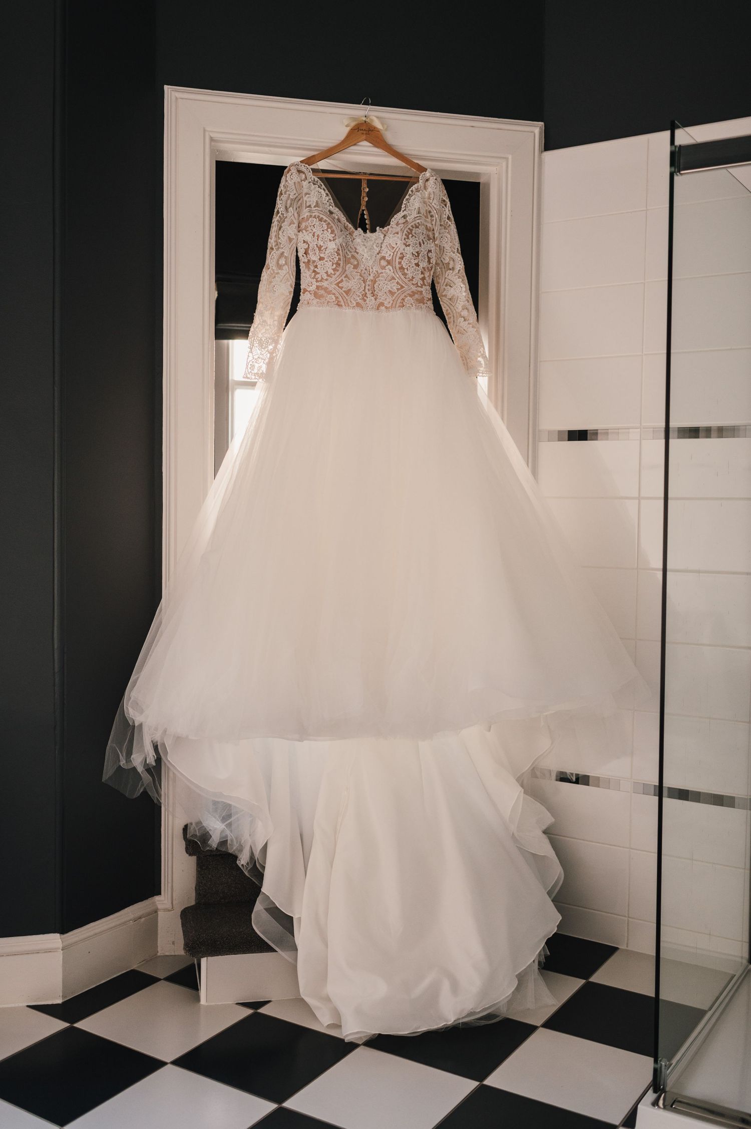 An elegant wedding gown with lace sleeves and tulle skirt displayed on a hanger in a bathroom with checkered floor.