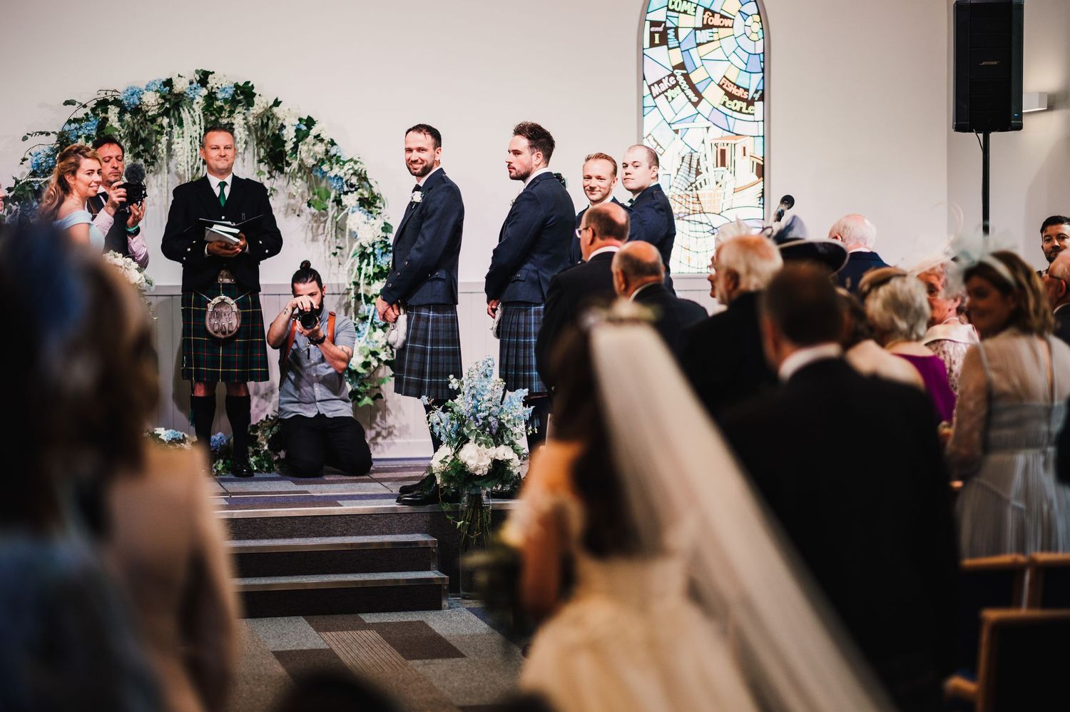 Wedding ceremony at church altar with floral arch and groomsmen in kilts standing in front.